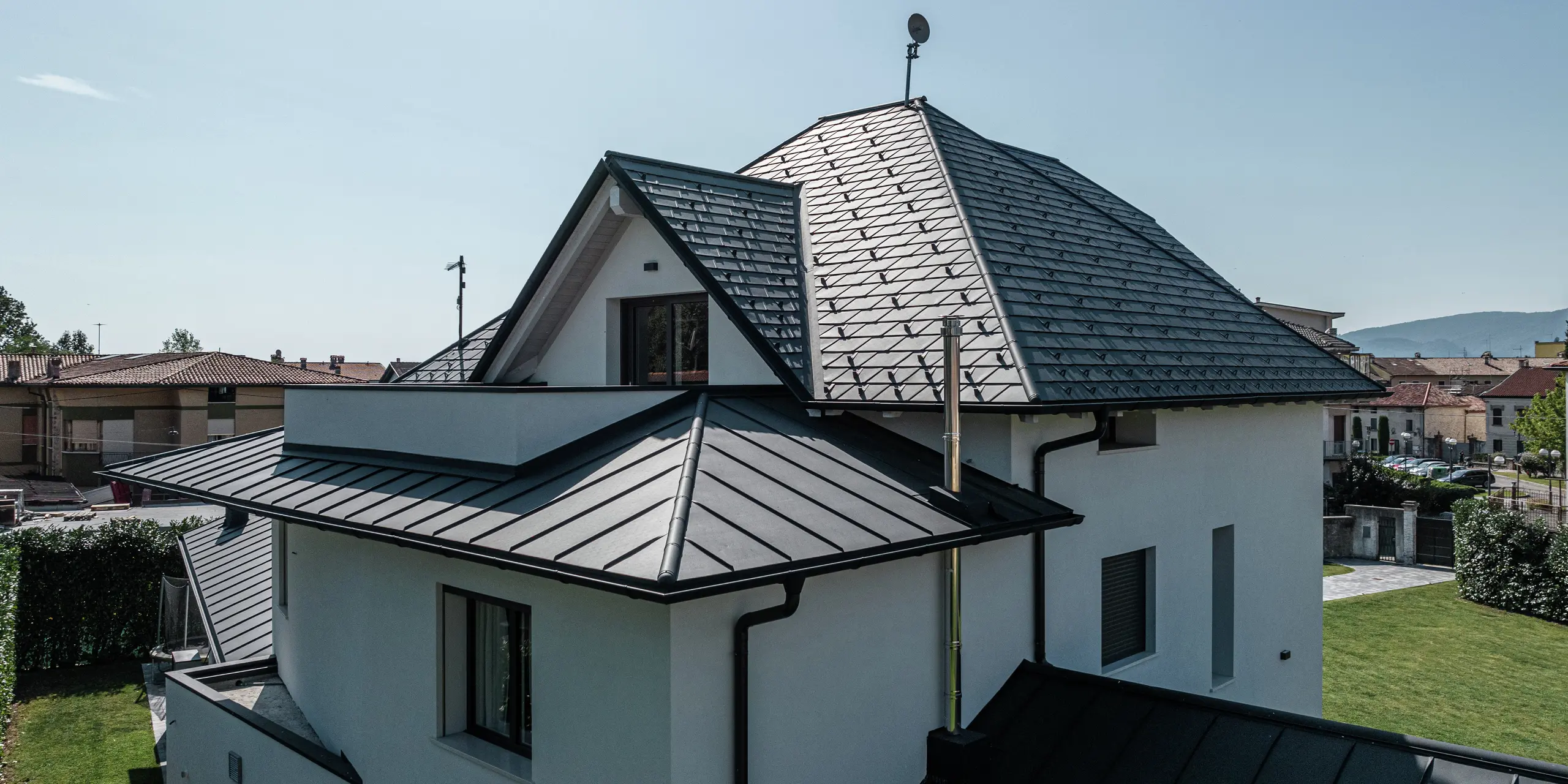 Vue détaillée du toit arrière d'une maison familiale moderne à Marano Vicentino, Italie. Le toit principal est recouvert de bardeaux PREFA en P.10 anthracite, tandis que les deux extensions sont équipées d'un élégant toit à double joint debout en aluminium. La combinaison de produits en aluminium de haute qualité garantit une solution de toiture durable, résistante aux intempéries et esthétique.