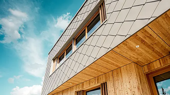 Le gros plan de la crèche d'Alzenau met en valeur la précision d'exécution et la qualité de l'association des losanges de façade 44x44 PREFA couleur bronze et du bois naturel. La structure diagonale de la façade en aluminium crée un motif d'écailles saisissant qui s'harmonise avec les persiennes verticales en bois entourant les fenêtres et le soffite en bois finement travaillé. La prise de vue en contre-plongée souligne la précision des lignes et le savoir-faire du bardage ventilé. Cette vue détaillée met en évidence la robustesse et la durabilité des produits PREFA, idéaux pour les projets architecturaux durables qui allient esthétique, fonctionnalité et responsabilité écologique au plus haut niveau.