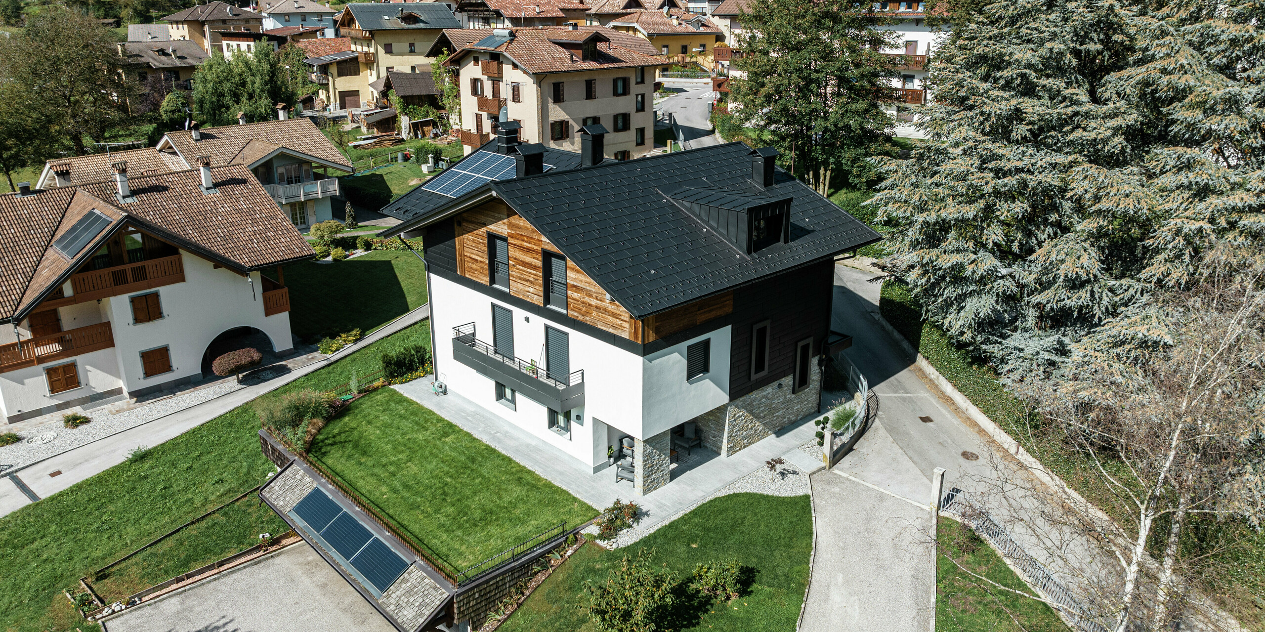 Vue d'un immeuble résidentiel rénové de façon moderne à Pelugo, en Italie, avec des R.16 PREFA en P.10 anthracite et des panneaux de façade PREFA Siding.X. La combinaison de l'aluminium, du bois et de la pierre assure une architecture harmonieuse, résistante aux intempéries et durable.