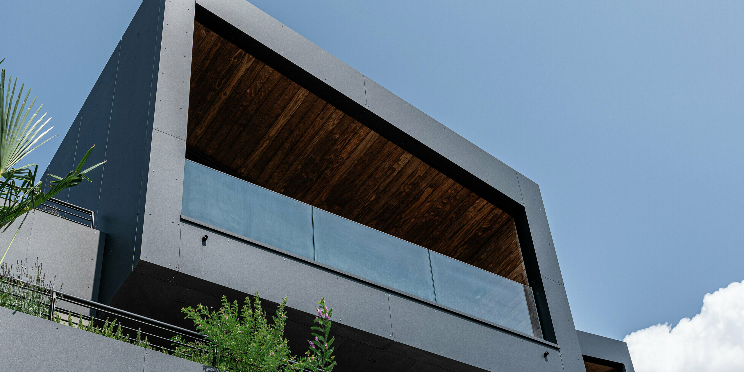 L'architecture moderne rencontre des matériaux durables : la forme cubique minimaliste avec un toit en pente et une terrasse couverte remarquable est parfaitement mise en valeur par la robuste façade en aluminium PREFA en P.10 Noir. Le dessous souligne les lignes claires et la durabilité du matériau, tandis que la verdure intégrée assure une harmonie naturelle. Résistante aux intempéries, facile à entretenir et élégante : l'enveloppe du bâtiment PREFA garantit protection et durabilité dans toutes les situations.