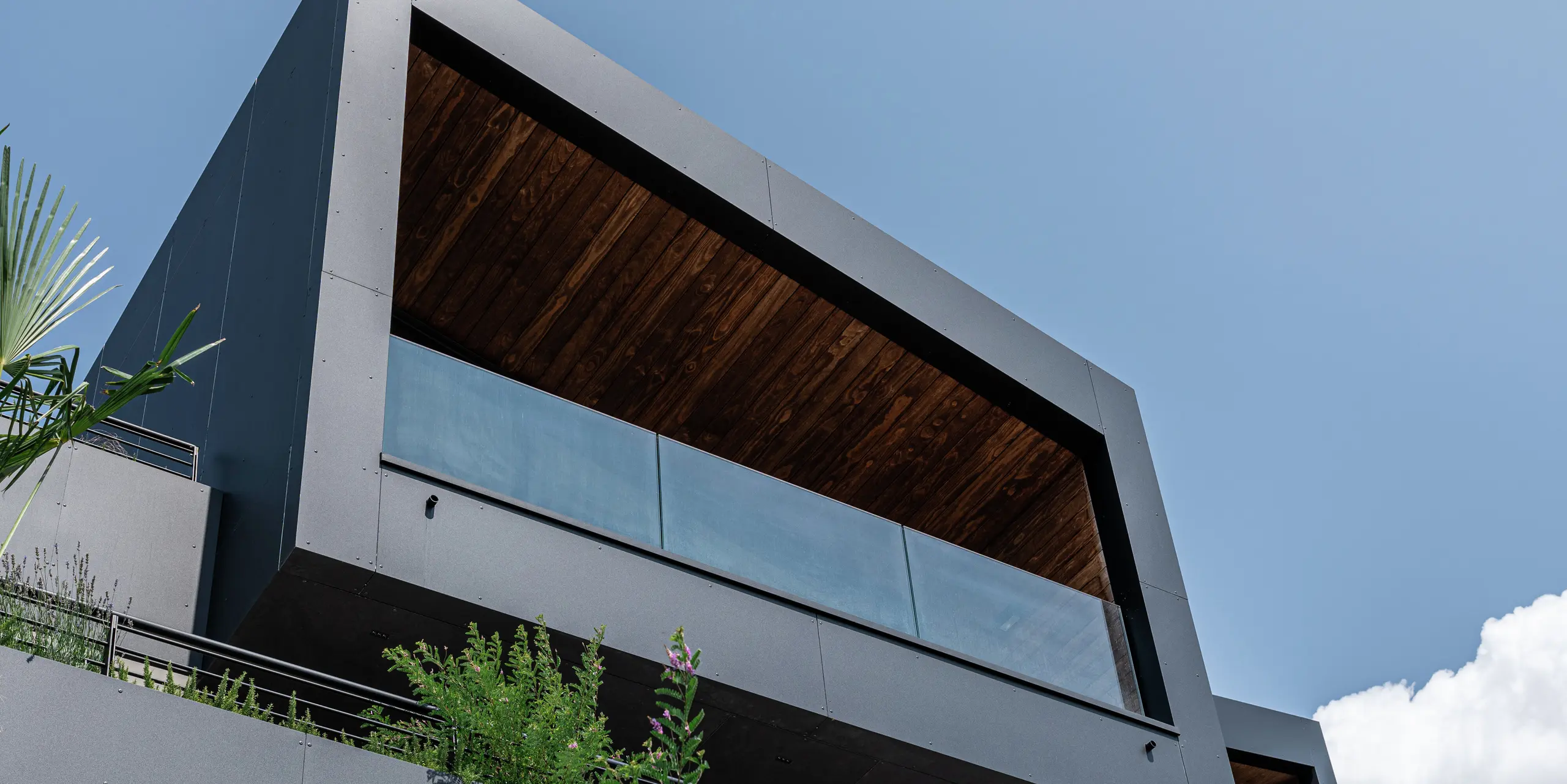 L'architecture moderne rencontre des matériaux durables : la forme cubique minimaliste avec un toit en pente et une terrasse couverte remarquable est parfaitement mise en valeur par la robuste façade en aluminium PREFA en P.10 Noir. Le dessous souligne les lignes claires et la durabilité du matériau, tandis que la verdure intégrée assure une harmonie naturelle. Résistante aux intempéries, facile à entretenir et élégante : l'enveloppe du bâtiment PREFA garantit protection et durabilité dans toutes les situations.
