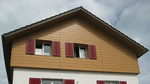 Habillage du pignon d’une maison classique avec toit à deux pans. Le pignon est habillé à l’aide de Sidings PREFA couleur chêne naturel posés à l’horizontale. Les fenêtres disposent de volets de couleur rouge.