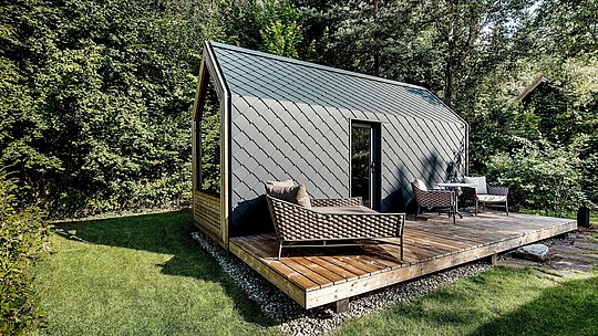 Une minuscule maison minimaliste, nichée au cœur du Secret Koi Garden sur les rives du lac Natterer, se dresse sur une vaste terrasse en bois, entourée d'une végétation forestière dense. Toit et façade sont revêtus de petits losanges en aluminium PREFA P.10 anthracite, dont la structure uniforme confère à la maison une allure résolument moderne. L'enveloppe en aluminium, robuste et résistante aux intempéries, offre une protection fiable et crée un contraste saisissant avec le bois, la verdure et l'espace de vie extérieur ouvert de ce complexe de glamping.