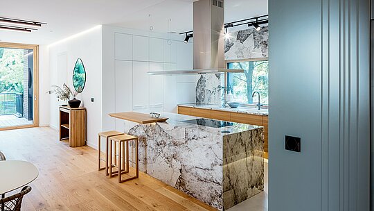 La photographie met en valeur l'élégant design intérieur d'une villa multigénérationnelle à Budapest, avec sa cuisine moderne ouverte. Parmi les éléments remarquables, on note les surfaces en pierre naturelle marbrée de l'îlot central et de la crédence, associées à des touches de chêne chaleureux et à des meubles intégrés de haute qualité. De larges fenêtres laissent entrer une abondante lumière naturelle, créant une harmonie entre l'espace et la verdure environnante. La pureté des lignes, le raffinement des matériaux et la précision de la réalisation témoignent du standing architectural du bâtiment, sublimé par la robustesse des produits en aluminium PREFA à l'extérieur.