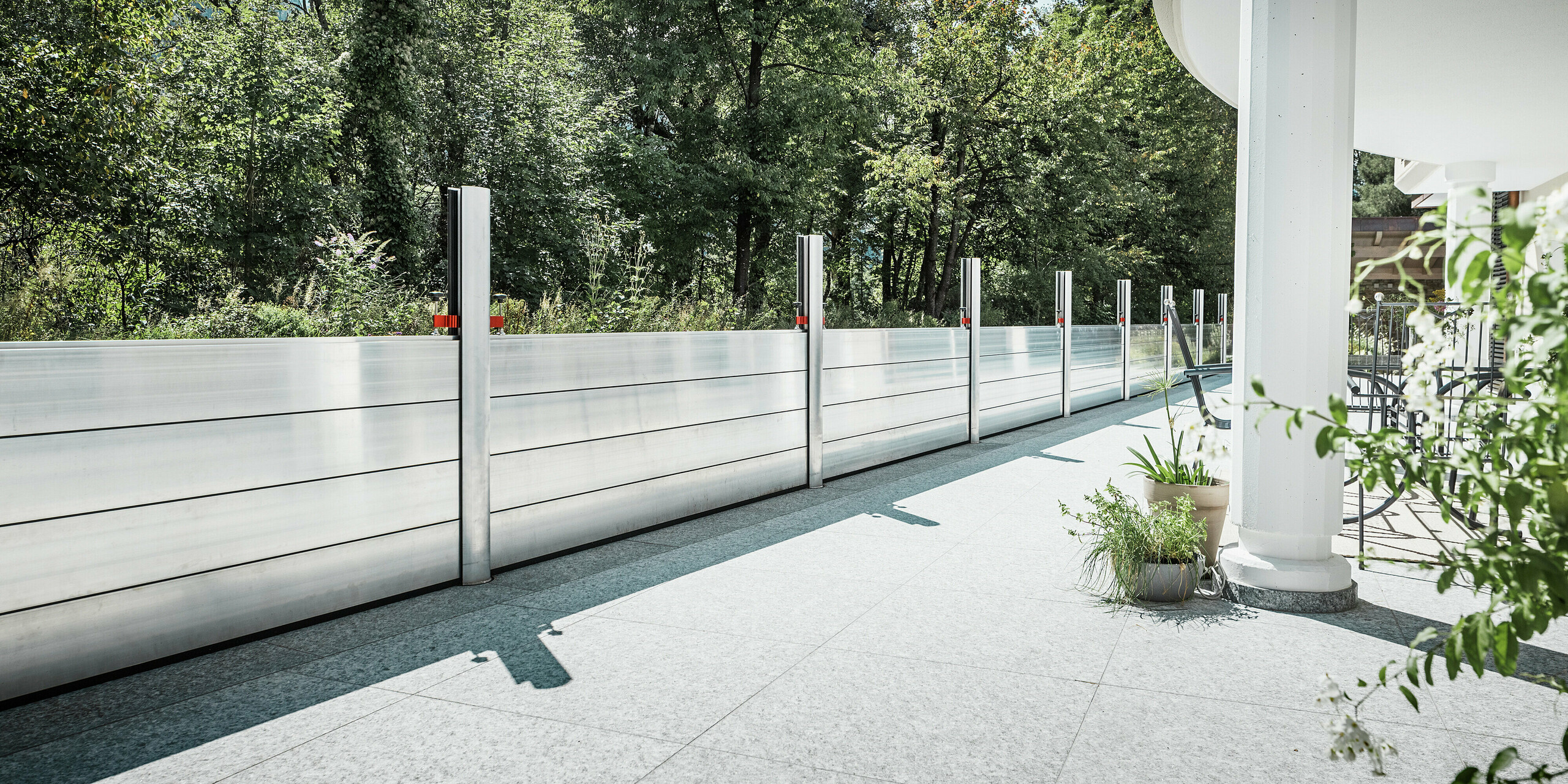 Vue depuis une élégante terrasse abritée, équipée d'un système de protection contre les crues PREFA. Les batardeaux en aluminium s'étendent en ligne droite le long de la limite de propriété jusqu'à la forêt adjacente, offrant une protection efficace contre les inondations. Les éléments en aluminium brillant s'intègrent harmonieusement au design moderne de la maison. Au premier plan : colonnes blanches, jardinières et mobilier de jardin, soulignant l'atmosphère chaleureuse.