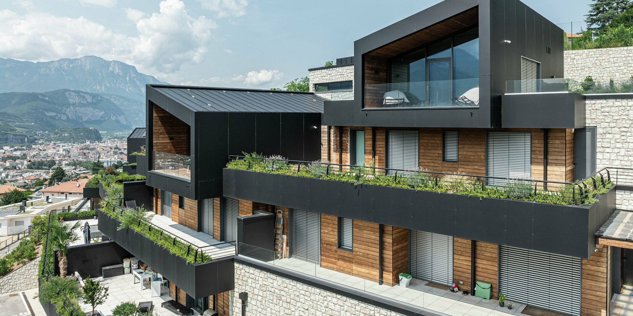 Architecture résidentielle moderne avec du caractère : L'élégante façade en panneaux composites en aluminium PREFA en noir P.10 s'harmonise parfaitement avec les éléments en bois chaleureux et la base en pierre naturelle. Le toit PREFALZ en P.10 Noir offre non seulement une unité esthétique mais également une résistance maximale aux intempéries. De généreuses surfaces vitrées et des terrasses vertes créent une connexion parfaite entre l'espace intérieur et extérieur, tandis que l'emplacement à flanc de colline offre une vue impressionnante sur Trente. Durable, facile d’entretien et élégant – un concept d’habitat qui a de l’avenir.