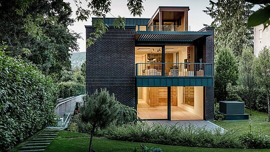 L'arrière de cette villa moderne et multigénérationnelle de Budapest est niché au cœur d'un jardin luxuriant. De larges baies vitrées ouvrent l'espace de vie sur l'extérieur, laissant pénétrer une douce lumière intérieure qui illumine la façade en briques texturées. Les lignes épurées du bâtiment sont mises en valeur par l'aluminium P.10 PREFALZ anthracite, utilisé pour le revêtement des balcons et certains éléments de façade, ce qui lui confère un attrait visuel durable. L'alliance de la brique, de l'aluminium et d'une végétation abondante souligne le caractère intemporel, raffiné et architecturalement sophistiqué de la maison.