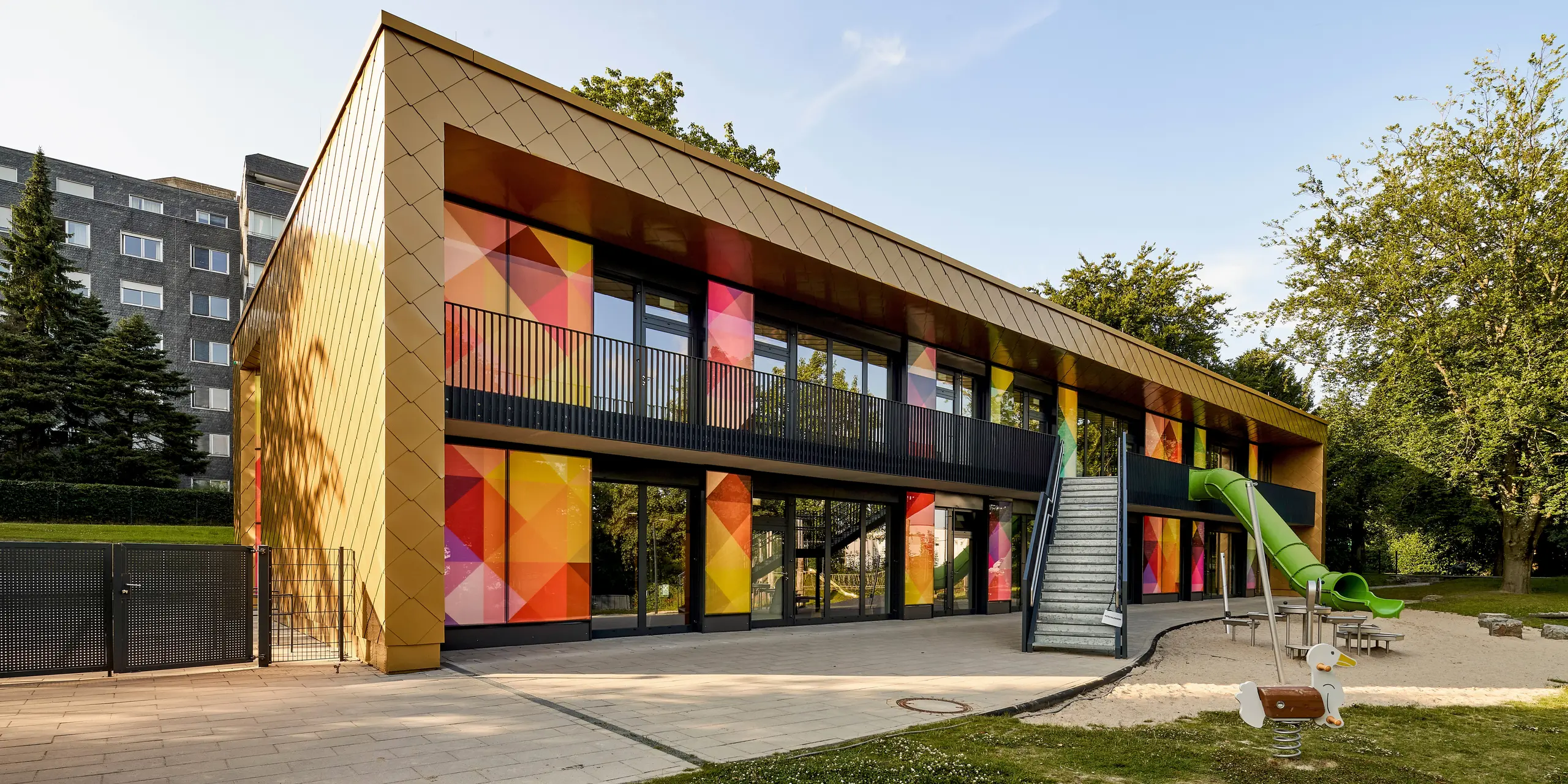 Ici, l'architecture contemporaine rencontre une spectaculaire façade en aluminium PREFA, ornée de panneaux muraux dorés en forme de losange. De larges surfaces vitrées aux motifs géométriques et aux couleurs intenses apportent des touches de dynamisme et créent des contrastes saisissants. L'entrée ouverte, le balcon et l'aire de jeux soulignent la vocation du bâtiment : une crèche. Les produits en aluminium PREFA impressionnent par leur extrême durabilité, leur résistance aux intempéries et leurs couleurs éclatantes. Ils constituent une solution durable et sans entretien pour des façades expressives d'une grande qualité esthétique.