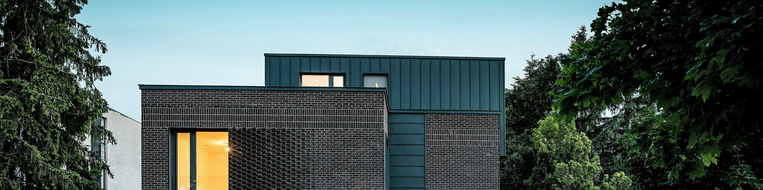 L'image présente un immeuble d'appartements moderne à Budapest, dont la toiture cubique, revêtue d'aluminium PREFALZ P.10 anthracite, contraste de façon saisissante avec le paysage historique environnant. Les joints verticaux créent un jeu de lumière captivant et soulignent la pureté des lignes architecturales. Les produits en aluminium PREFA, de haute qualité, confèrent au bâtiment une élégance intemporelle, tout en impressionnant par leur durabilité, leur résistance aux intempéries et leur finition impeccable. La façade et la toiture anthracite attirent immédiatement le regard et mettent en valeur le caractère novateur du bâtiment.