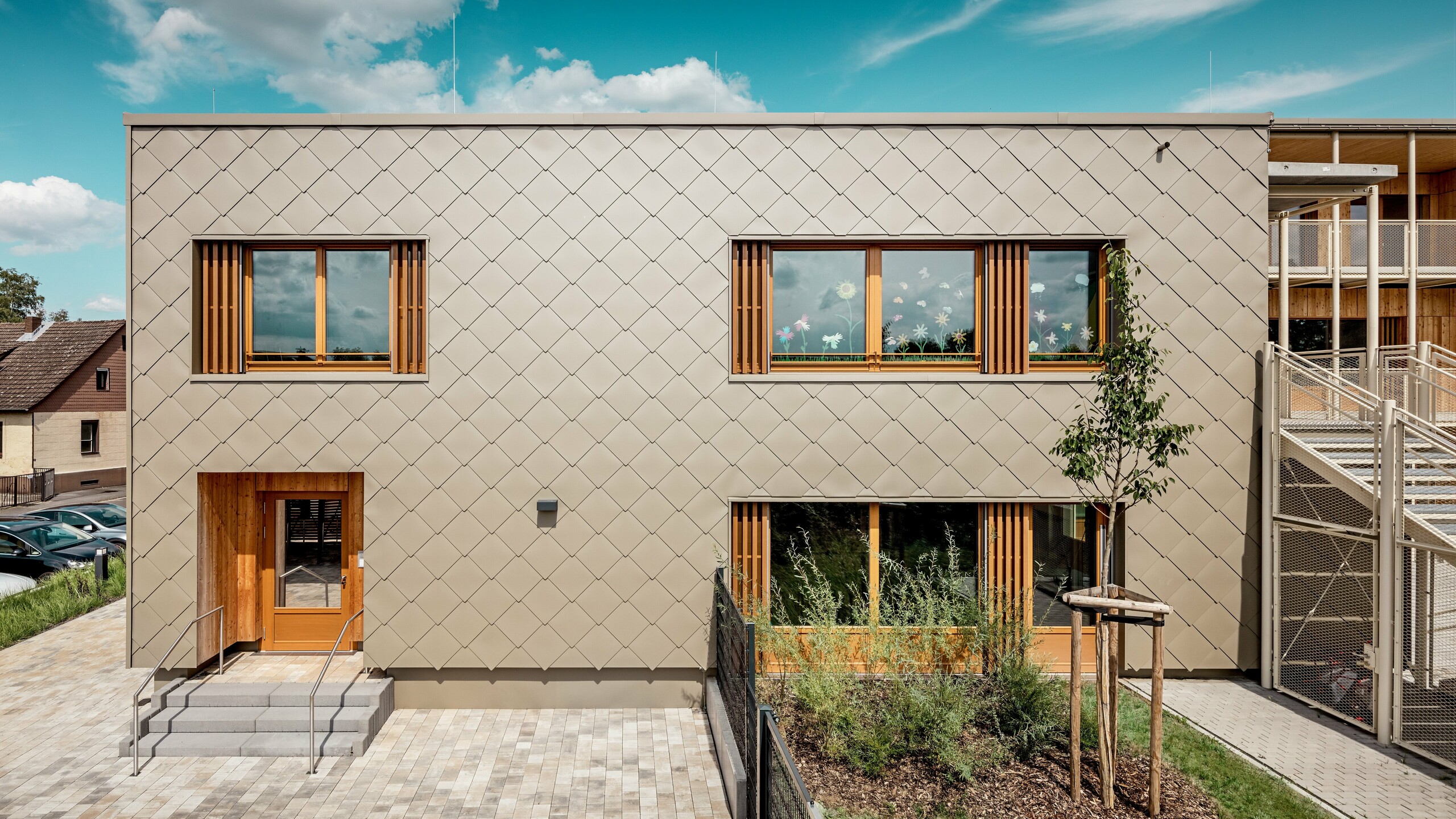 La façade arrière de la crèche moderne d'Alzenau, vue de face, impressionne par ses lignes épurées et son élégante façade composée de losanges de façade 44x44 PREFA couleur bronze. La structure géométrique de la façade crée un motif harmonieux, évoquant des écailles, accentué par l'horizontalité apaisante des fenêtres. De généreux encadrements en bois et des persiennes verticales apportent une touche naturelle et créent une atmosphère chaleureuse et accueillante. L'alliance judicieuse de l'aluminium durable et du bois local témoigne d'une architecture durable et d'une grande qualité de conception. L'image met en lumière la précision de l'exécution, le choix judicieux des matériaux et l'intégration réussie du bâtiment dans son environnement urbain.