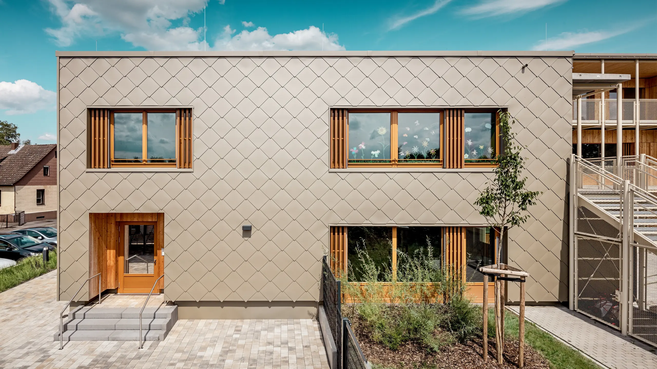 La façade arrière de la crèche moderne d'Alzenau, vue de face, impressionne par ses lignes épurées et son élégante façade composée de losanges de façade 44x44 PREFA couleur bronze. La structure géométrique de la façade crée un motif harmonieux, évoquant des écailles, accentué par l'horizontalité apaisante des fenêtres. De généreux encadrements en bois et des persiennes verticales apportent une touche naturelle et créent une atmosphère chaleureuse et accueillante. L'alliance judicieuse de l'aluminium durable et du bois local témoigne d'une architecture durable et d'une grande qualité de conception. L'image met en lumière la précision de l'exécution, le choix judicieux des matériaux et l'intégration réussie du bâtiment dans son environnement urbain.