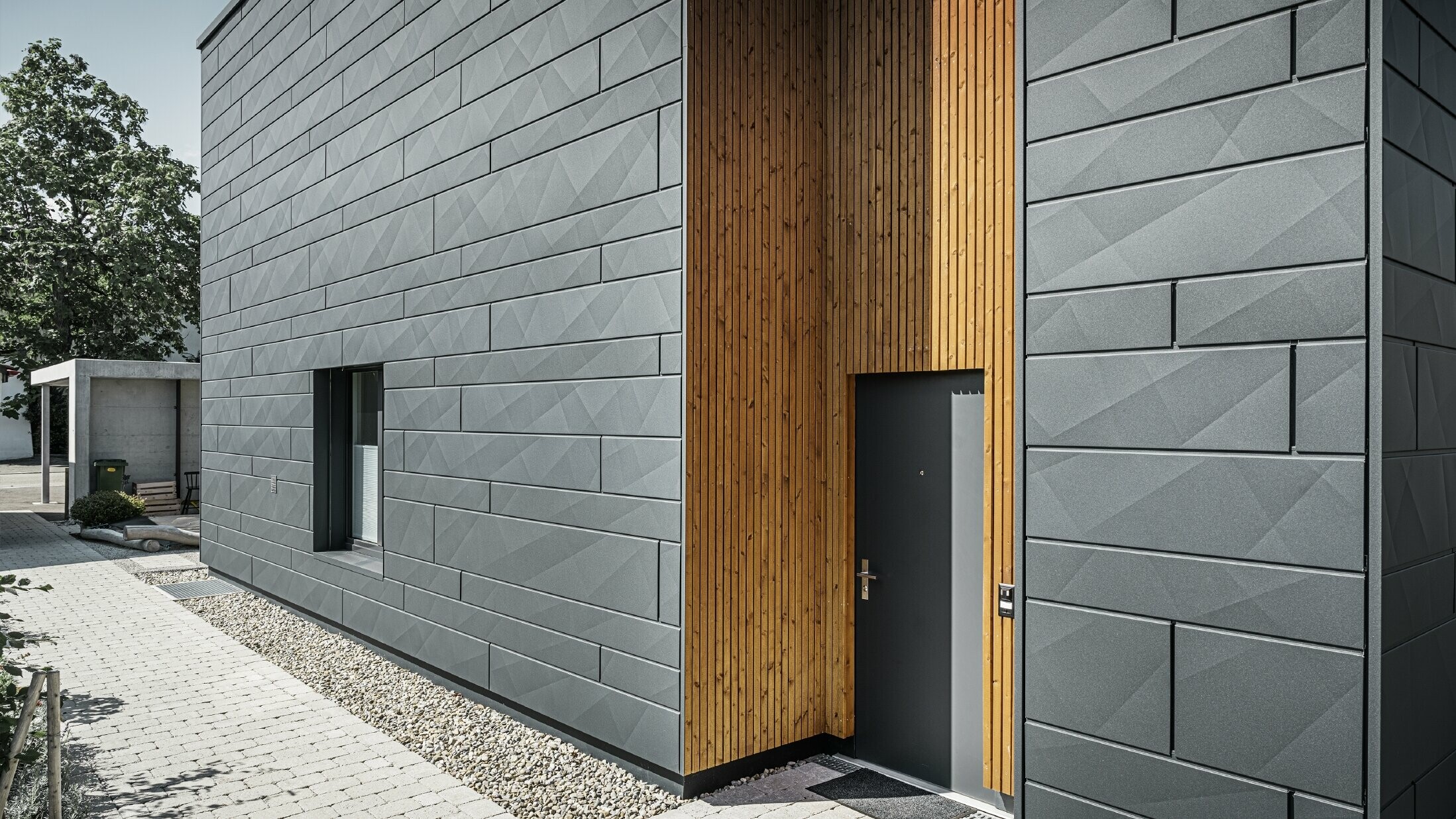 On peut voir l’entrée de la maison individuelle moderne. La façade est habillée de Siding.X PREFA, la zone autour de la porte est en bois.