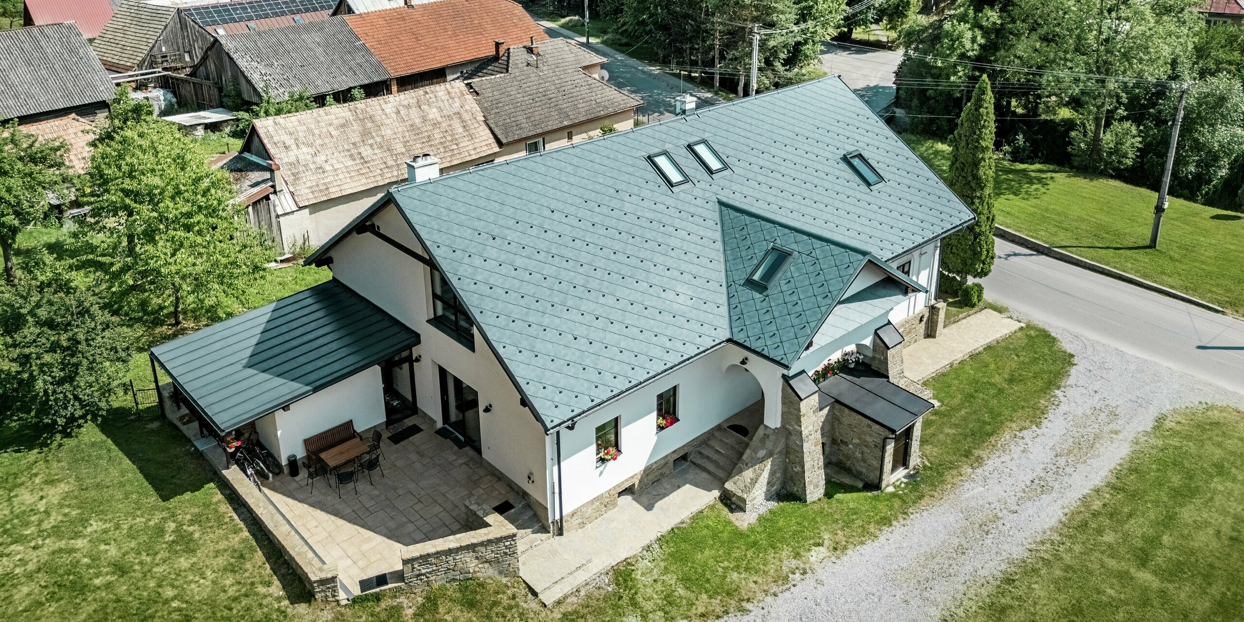 L'image montre une maison d'artisan à Klin, en Slovaquie, vue du ciel. Le toit à deux pans, recouvert de losanges PREFA 44 x 44 couleur Gris Pierre P.10, s'étend avec élégance au-dessus du bâtiment et s'intègre harmonieusement à son environnement. La disposition symétrique des pans de toiture et les puits de lumière intégrés assurent un éclairage homogène à l'intérieur. Le style moderne de la toiture contraste parfaitement avec l'architecture traditionnelle de la maison, caractérisée par sa maçonnerie en pierre naturelle et ses façades blanches. La toiture en aluminium est non seulement un atout esthétique, mais aussi très fonctionnelle : légère, durable et résistante à la corrosion, elle est idéale pour la rénovation de bâtiments historiques.
