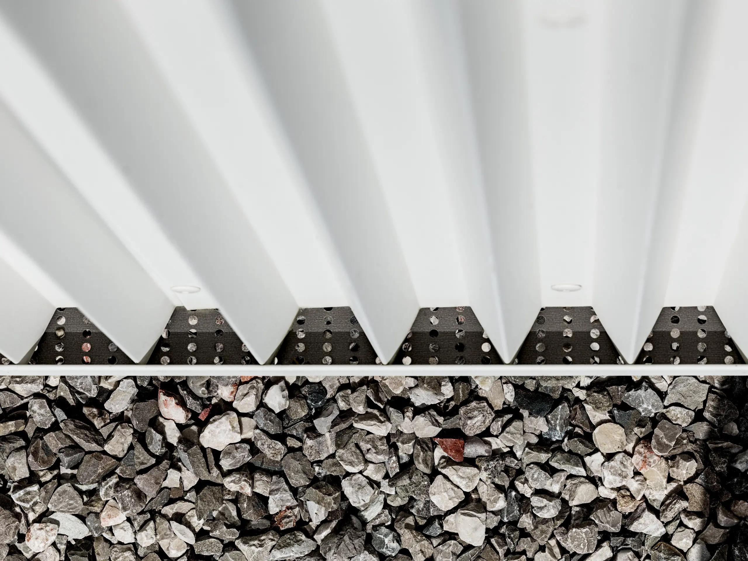 L'image présente un détail de la façade ventilée PREFALZ en aluminium blanc pur, caractérisée par des formes en zigzag pliées avec précision. Cette technique de pliage innovante garantit non seulement une structure esthétique, mais aussi une excellente ventilation et une grande résistance aux intempéries. Les éléments de façade sont assemblés à l'aide de fixations invisibles spécifiques, ce qui confère à la surface une apparence particulièrement propre et durable. La partie inférieure de l'image montre le revêtement de gravier entourant le bâtiment, assurant le drainage et la stabilisation, et soulignant ainsi la fonctionnalité de la conception de la façade. L'utilisation de PREFALZ répond aux exigences esthétiques et techniques, ce qui en fait un choix idéal pour une architecture moderne et durable.
