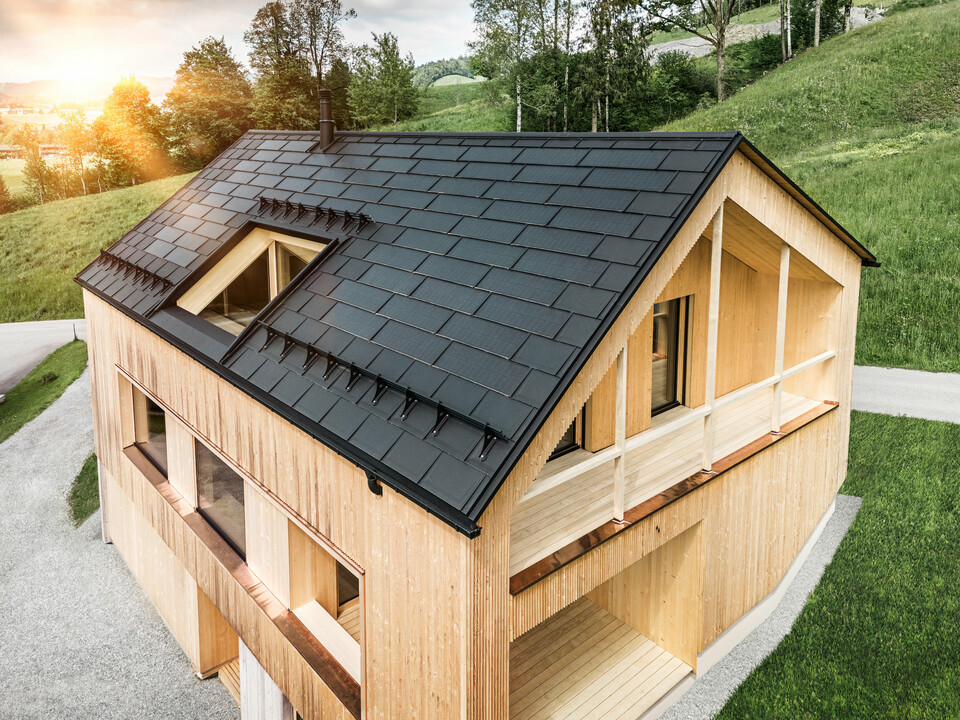 Prise de vue d'une maison en bois moderne et durable à Egg avec un toit solaire PREFA, qui se distingue par son aspect sombre, structuré et élégant. Les modules solaires sont stratégiquement placés entre les R.16 classiques pour profiter d'un rayonnement solaire optimal et s'intégrer esthétiquement dans la conception globale. La maison est en harmonie avec son environnement verdoyant, tandis que le chaud soleil du soir met en valeur la texture naturelle du bois de la façade et souligne la solidité de la construction. Un exemple harmonieux de bâtiment respectueux de l’environnement et d’efficacité énergétique.