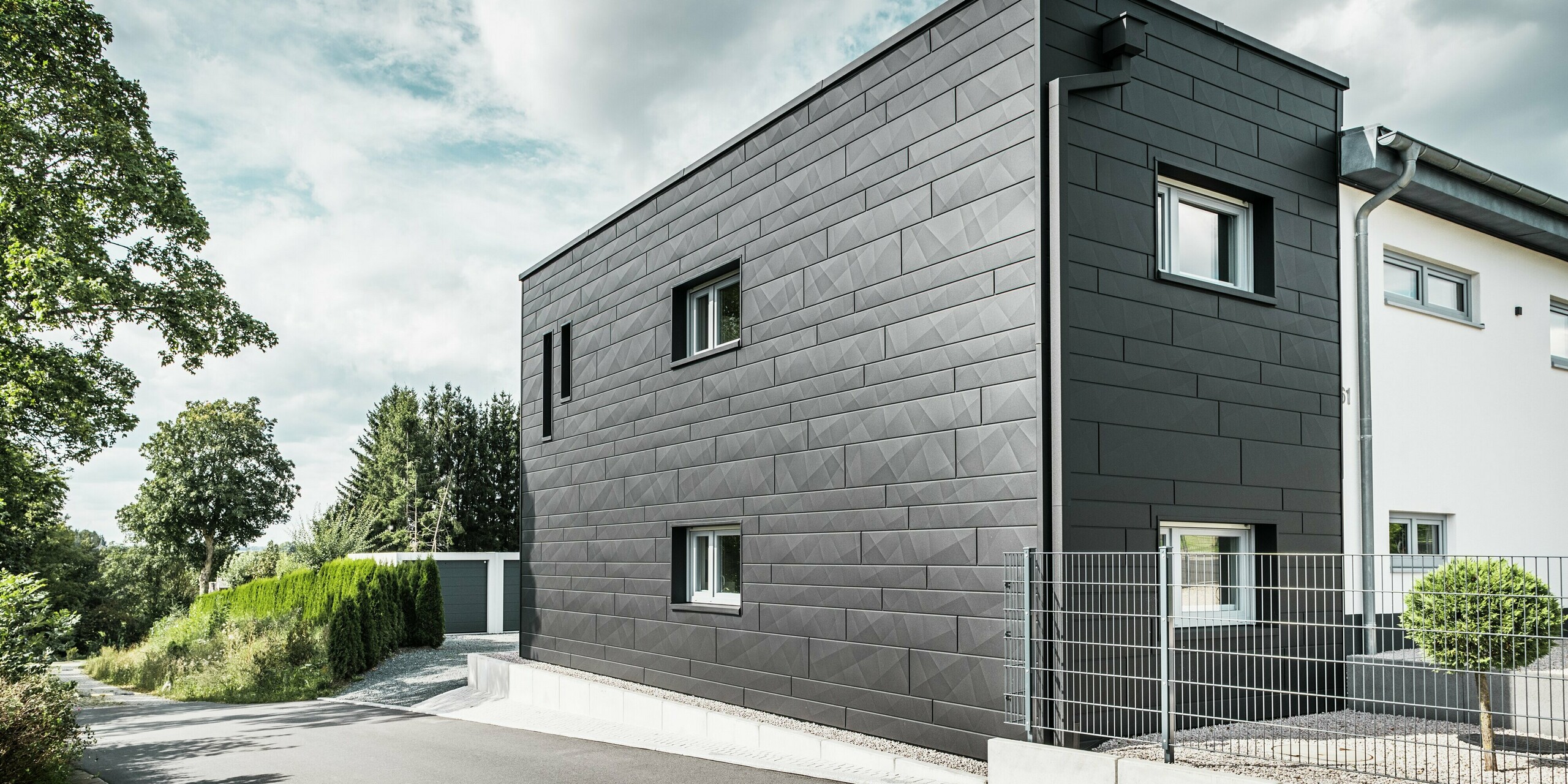 L'arrière de la maison individuelle modernisée de Teuschnitz impressionne par son élégante façade en Siding.X PREFA couleur P.10 noir. Les panneaux en aluminium géométriques créent un aspect structuré et saisissant et confèrent au bâtiment un caractère moderne et intemporel. Le système intégré de gouttière PREFA, avec tuyau carré et boîte à eau, s'intègre harmonieusement à l'ensemble, alliant esthétique et fonctionnalité.