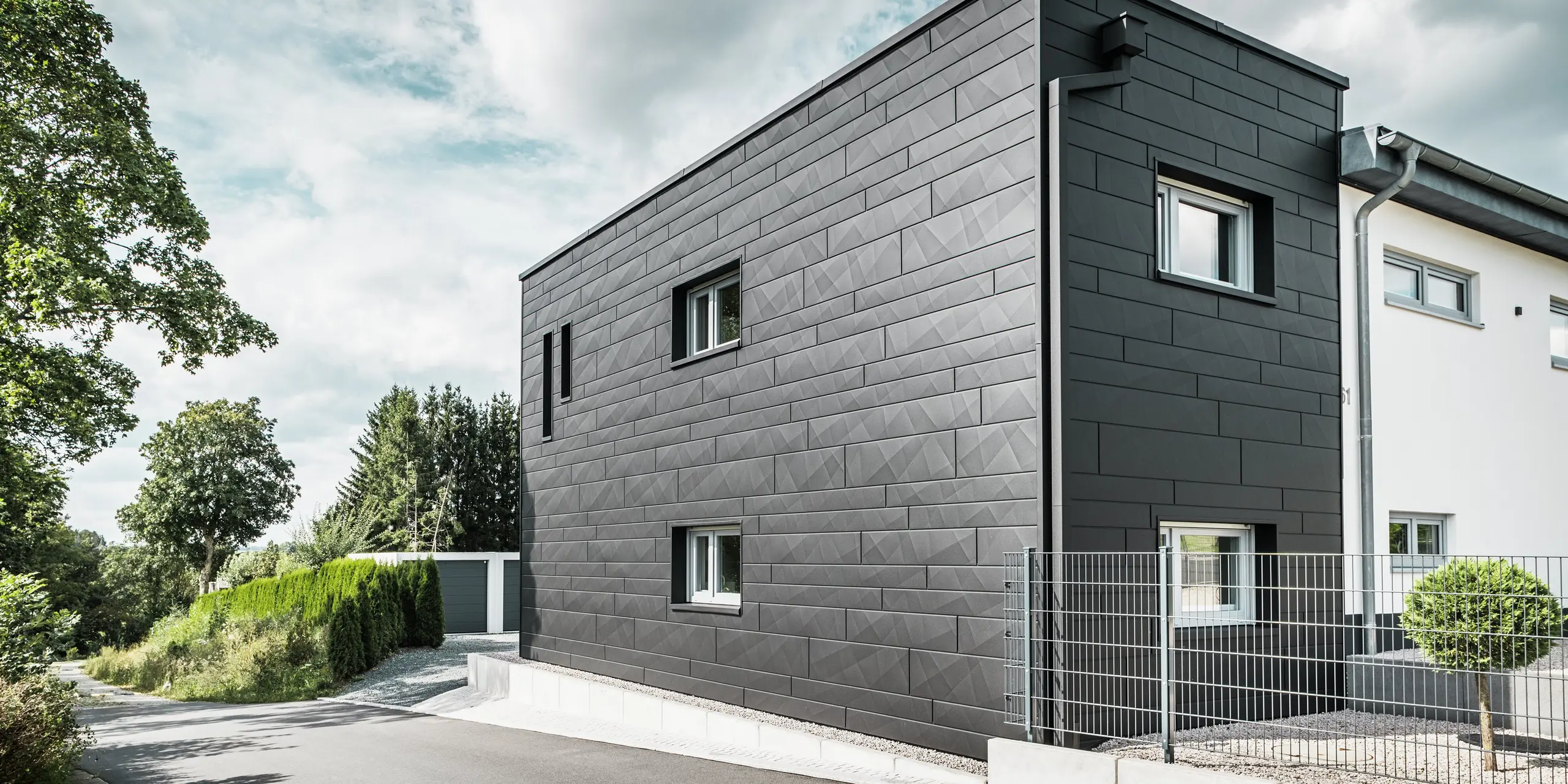L'arrière de la maison individuelle modernisée de Teuschnitz impressionne par son élégante façade en Siding.X PREFA couleur P.10 noir. Les panneaux en aluminium géométriques créent un aspect structuré et saisissant et confèrent au bâtiment un caractère moderne et intemporel. Le système intégré de gouttière PREFA, avec tuyau carré et boîte à eau, s'intègre harmonieusement à l'ensemble, alliant esthétique et fonctionnalité.