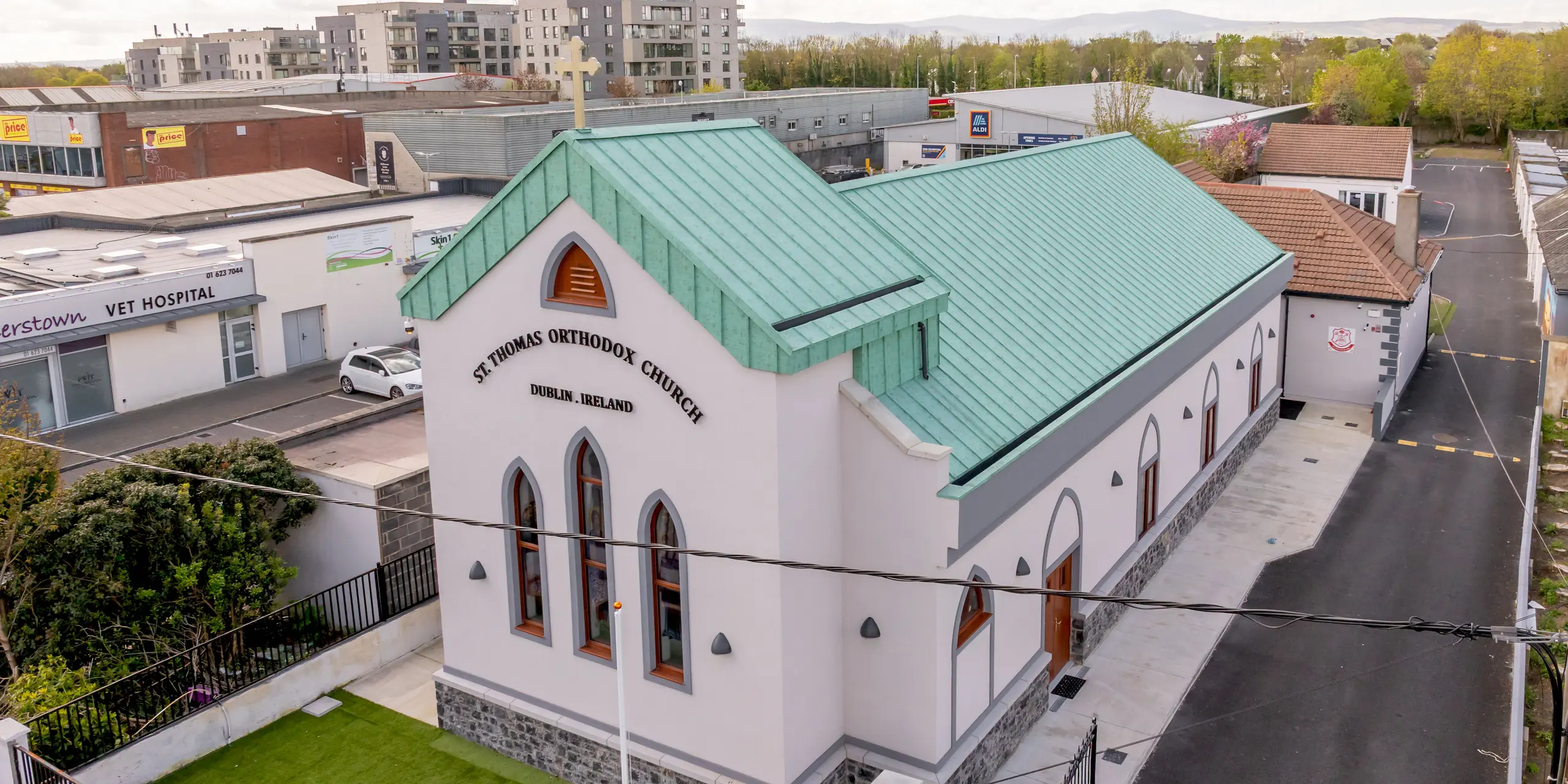 Images aériennes de l'église orthodoxe Saint-Thomas de Palmersgate, à Dublin, révélant son impressionnante toiture à joints debout en aluminium PREFALZ, couleur P.10 vert de gris. Cette vaste toiture structure l'architecture de l'édifice et, grâce à ses joints précis, sa croix imposante et ses fenêtres cintrées caractéristiques, constitue un point focal remarquable. La teinte vert patiné rappelle les toitures en cuivre traditionnelles et confère à l'église une élégance intemporelle. L'aluminium PREFALZ, de haute qualité, se distingue par sa durabilité, sa résistance à la corrosion, sa légèreté et son caractère écologique. Ainsi, la toiture PREFALZ allie harmonieusement technologie moderne et architecture traditionnelle.