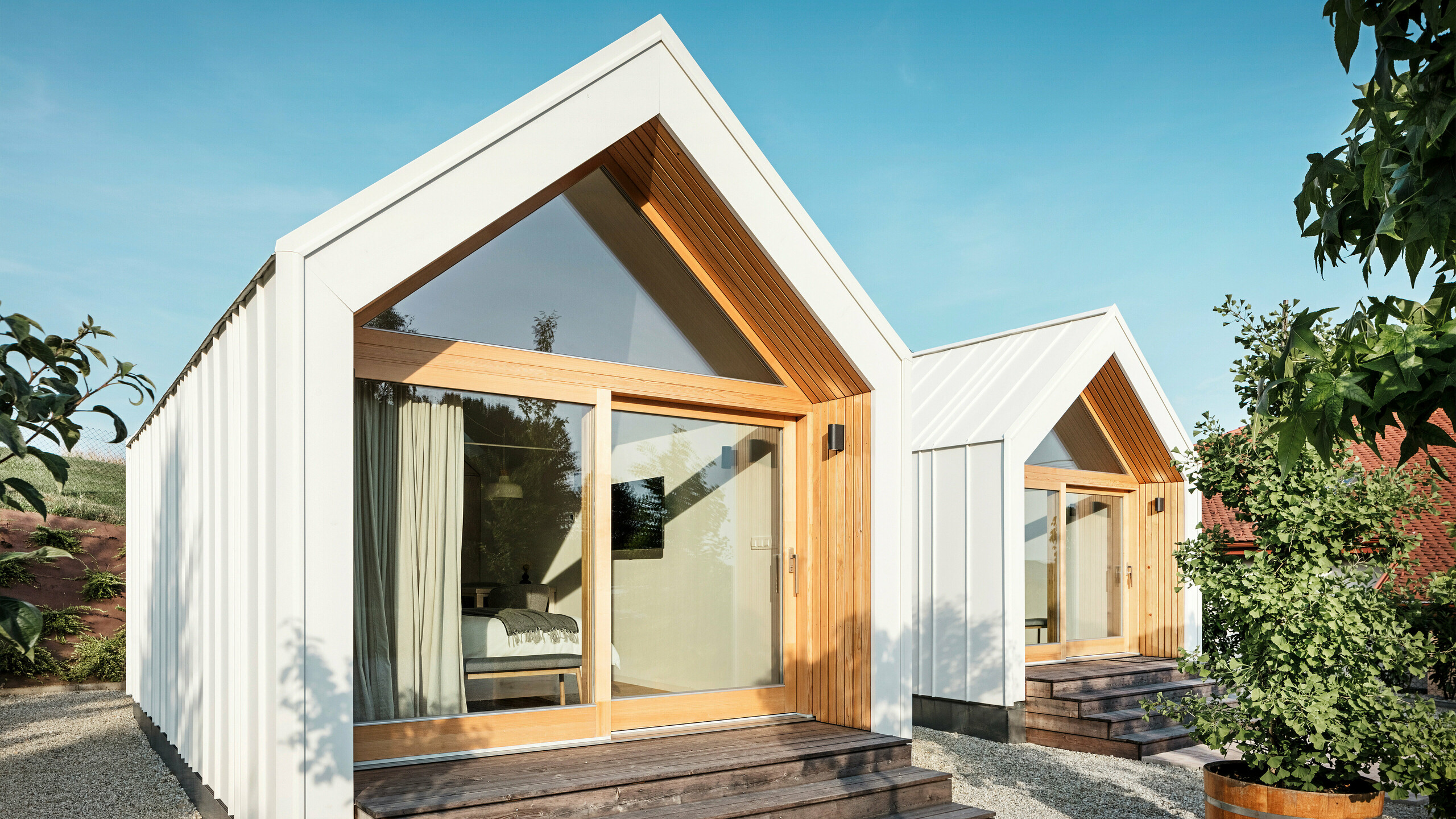 Vue de face des deux petites maisons de la résidence d'hôtes « Pri Momi » à Kančevci, en Slovénie, avec de grandes façades vitrées, conçues avec un toit et un revêtement de façade PREFALZ en blanc pur P.10 et bois naturel. Les micro-maisons minimalistes comportent de larges marches en bois menant aux entrées, offrant une vue directe sur les intérieurs confortables. Les lignes claires et la combinaison de l'aluminium et du bois soulignent le design moderne et accueillant du logement de vacances.