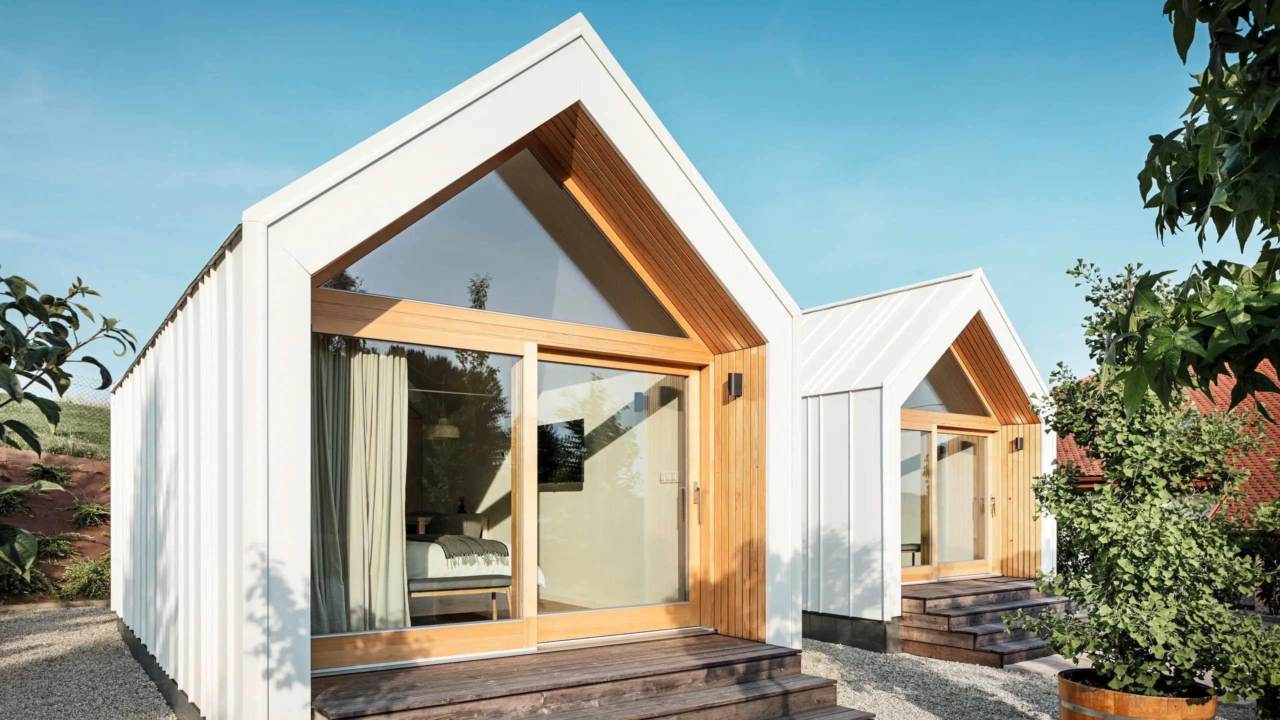 Vue de face des deux petites maisons de la résidence d'hôtes « Pri Momi » à Kančevci, en Slovénie, avec de grandes façades vitrées, conçues avec un toit et un revêtement de façade PREFALZ en blanc pur P.10 et bois naturel. Les micro-maisons minimalistes comportent de larges marches en bois menant aux entrées, offrant une vue directe sur les intérieurs confortables. Les lignes claires et la combinaison de l'aluminium et du bois soulignent le design moderne et accueillant du logement de vacances.