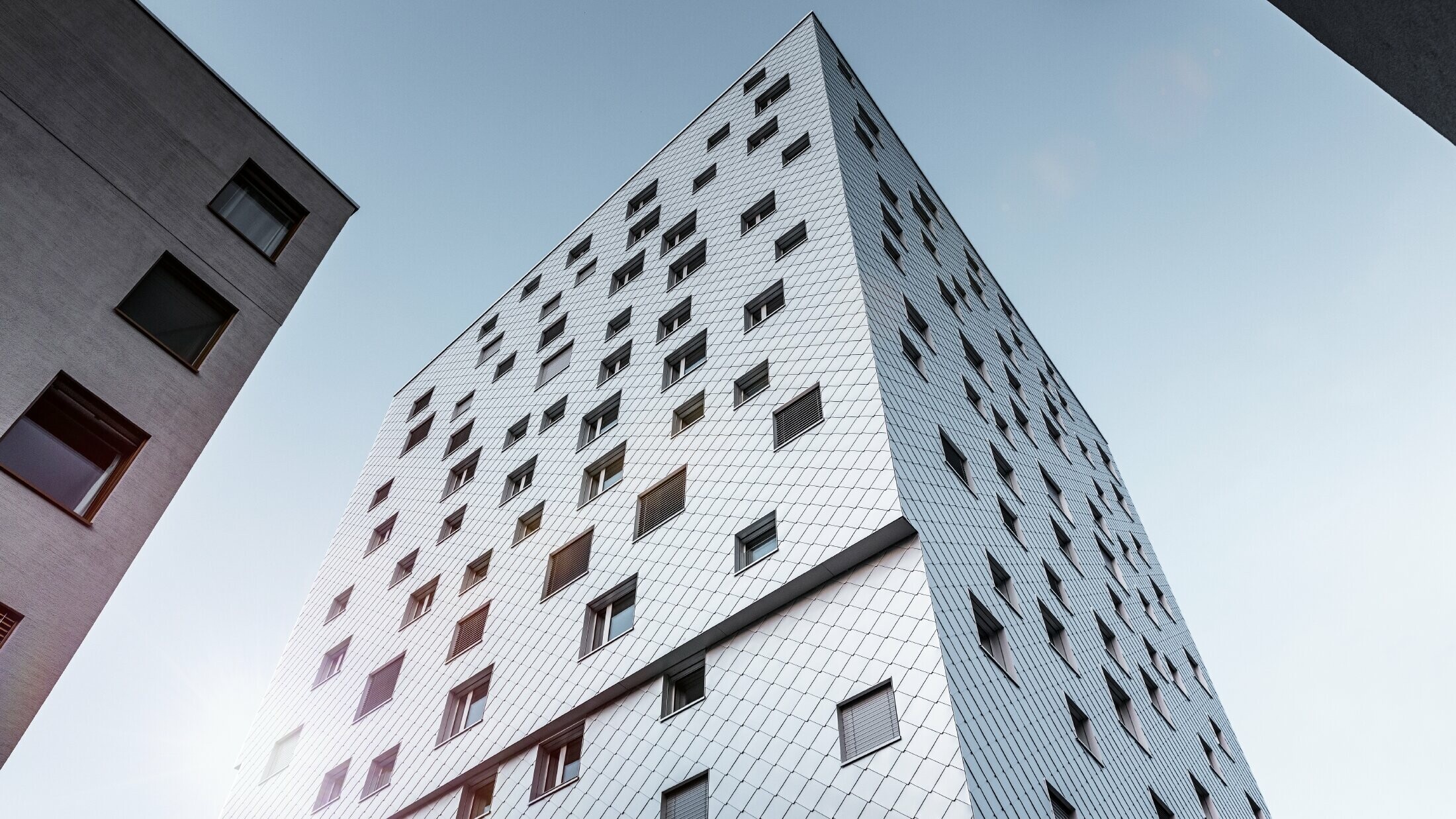 Das Bild zeigt eine Perspektive von unten auf das Solitaire-Hochhaus in Horw, Luzern, das von der Architektin Tilla Theus entworfen wurde. Die Fassade des Gebäudes ist mit eloxierten Aluminiumrauten von PREFA verkleidet, die ein diagonales Muster bilden und die Umgebung reflektieren. Die Fenster sind unregelmäßig angeordnet, was zu der charakteristischen Optik des Gebäudes beiträgt. Im Hintergrund sind Teile anderer Gebäude sichtbar, und der Himmel ist klar und blau.