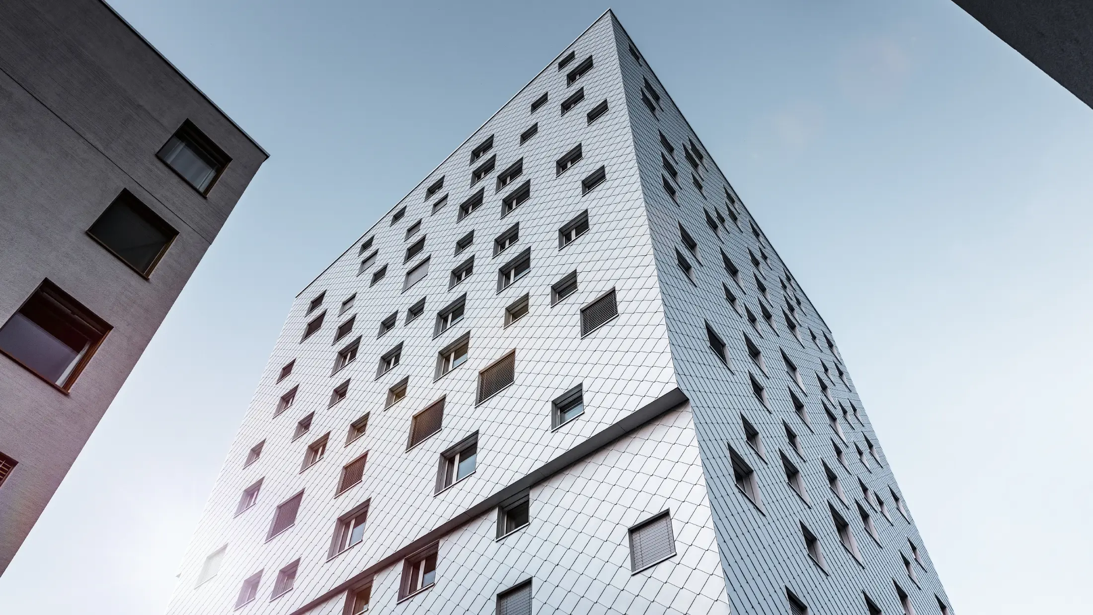 Das Bild zeigt eine Perspektive von unten auf das Solitaire-Hochhaus in Horw, Luzern, das von der Architektin Tilla Theus entworfen wurde. Die Fassade des Gebäudes ist mit eloxierten Aluminiumrauten von PREFA verkleidet, die ein diagonales Muster bilden und die Umgebung reflektieren. Die Fenster sind unregelmäßig angeordnet, was zu der charakteristischen Optik des Gebäudes beiträgt. Im Hintergrund sind Teile anderer Gebäude sichtbar, und der Himmel ist klar und blau.