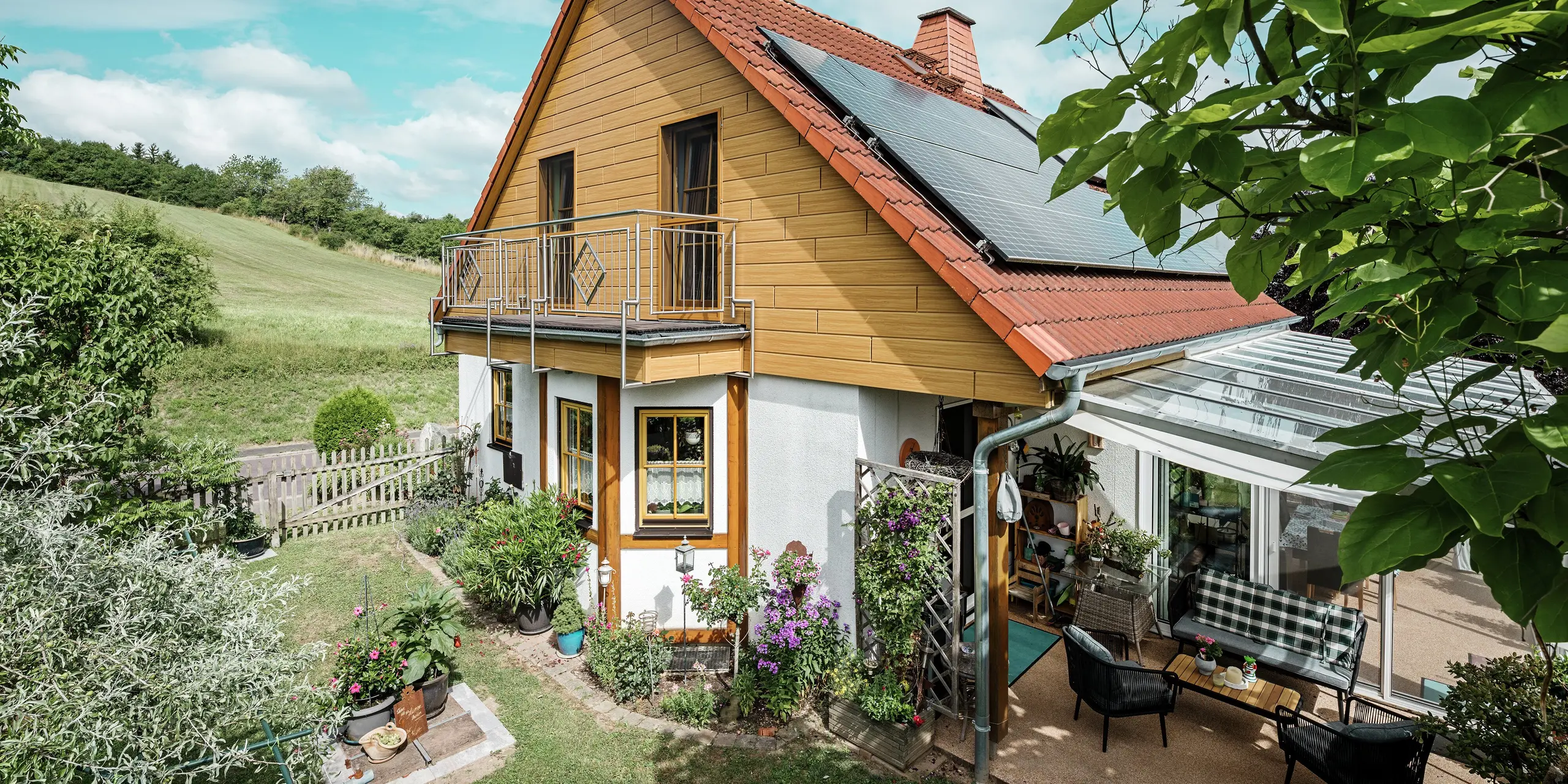La photo montre une charmante maison individuelle à Blieskastel, dont le pignon fraîchement rénové avec des Sidings PREFA couleur chêne naturel, attire immédiatement le regard. L'aspect bois de l'aluminium, plus durable, s'harmonise parfaitement avec le toit en tuiles rouges et la vaste véranda. La verdure luxuriante et le parterre de fleurs soigneusement aménagé forment une scène idyllique et vivante. La rénovation de façades avec PREFA confère à la maison un aspect naturel et en impose par sa robustesse : résistance aux intempéries, fidélité aux couleurs d'origine et longue durée de vie, pratiquement sans entretien.