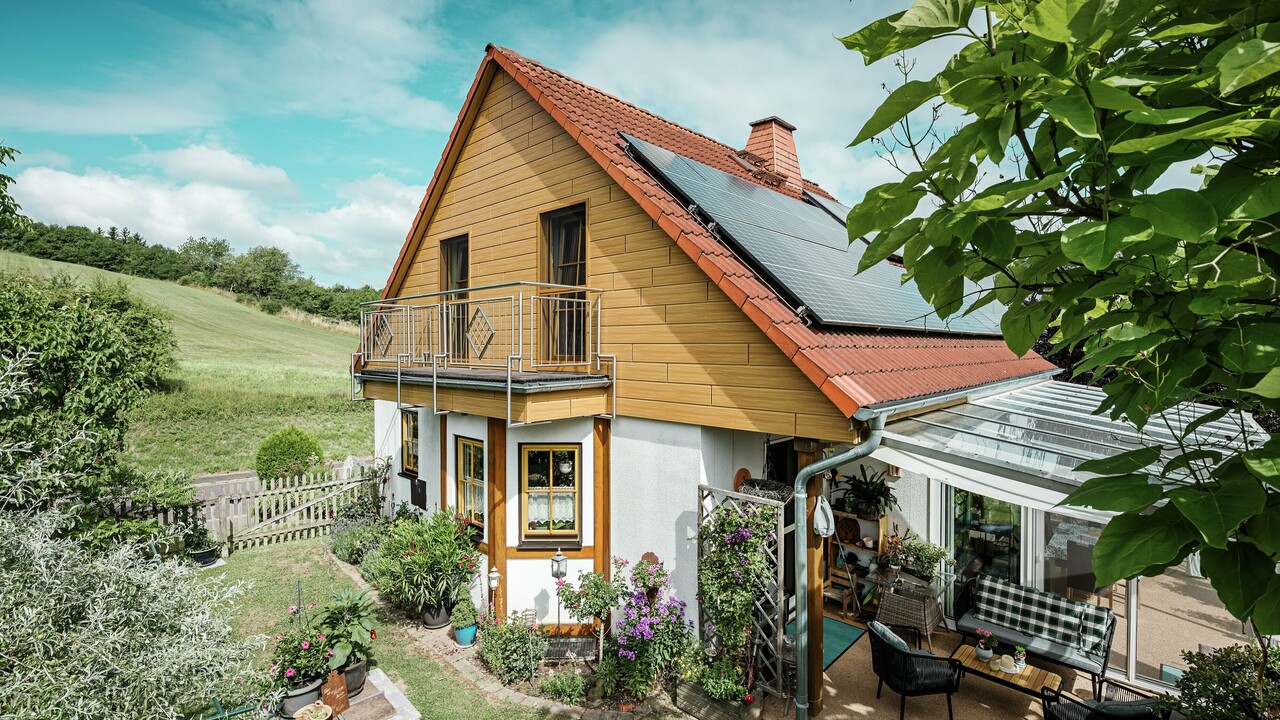 La photo montre une charmante maison individuelle à Blieskastel, dont le pignon fraîchement rénové avec des Sidings PREFA couleur chêne naturel, attire immédiatement le regard. L'aspect bois de l'aluminium, plus durable, s'harmonise parfaitement avec le toit en tuiles rouges et la vaste véranda. La verdure luxuriante et le parterre de fleurs soigneusement aménagé forment une scène idyllique et vivante. La rénovation de façades avec PREFA confère à la maison un aspect naturel et en impose par sa robustesse : résistance aux intempéries, fidélité aux couleurs d'origine et longue durée de vie, pratiquement sans entretien.