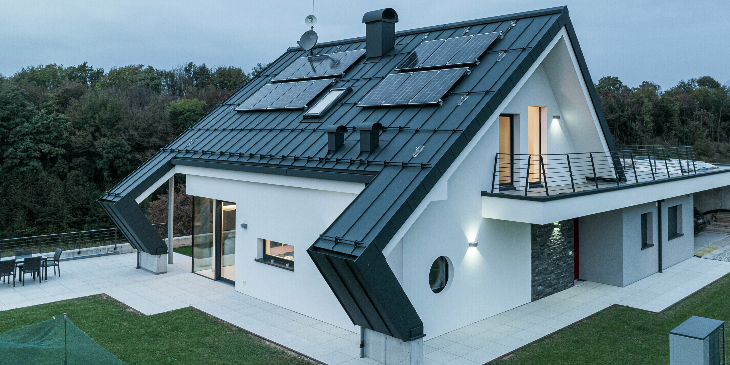 Maison individuelle moderne à Borgo Valbelluna avec toiture à joint debout PREFALZ en P.10 anthracite. Le toit en aluminium avec ses modules photovoltaïques allie production d'énergie durable et esthétique intemporelle. Les surplombs de toit marquants soulignent le design innovant et offrent une protection supplémentaire contre les intempéries. De grandes surfaces vitrées, une façade enduite de blanc et un balcon enveloppant créent une image globale harmonieuse.
