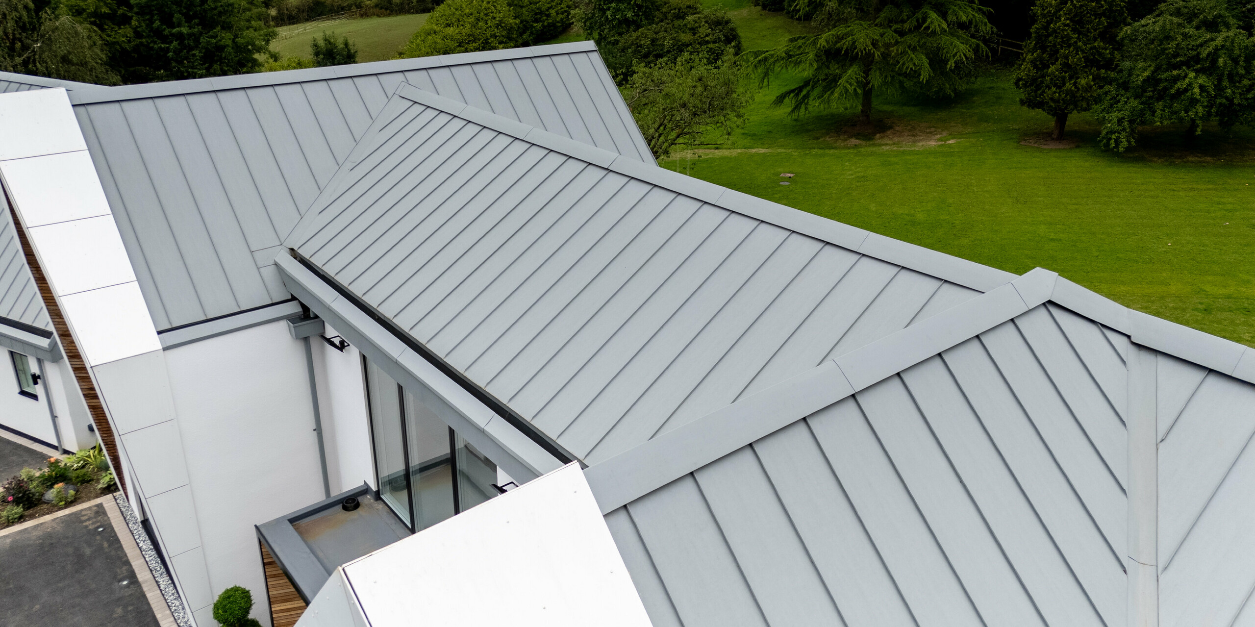 La photographie présente la faîtière de la toiture PREFALZ, réalisée avec précision et de couleur gris quartz, sous un angle unique. La toiture à joints debout, avec ses lignes épurées et son élégante surface métallique, se détache magnifiquement sur le paysage verdoyant. L'utilisation de PREFALZ garantit non seulement une esthétique soignée, mais aussi une grande durabilité et une excellente résistance aux intempéries. Son intégration parfaite au design moderne du bâtiment illustre avec brio la fusion harmonieuse de la forme et de la fonction.