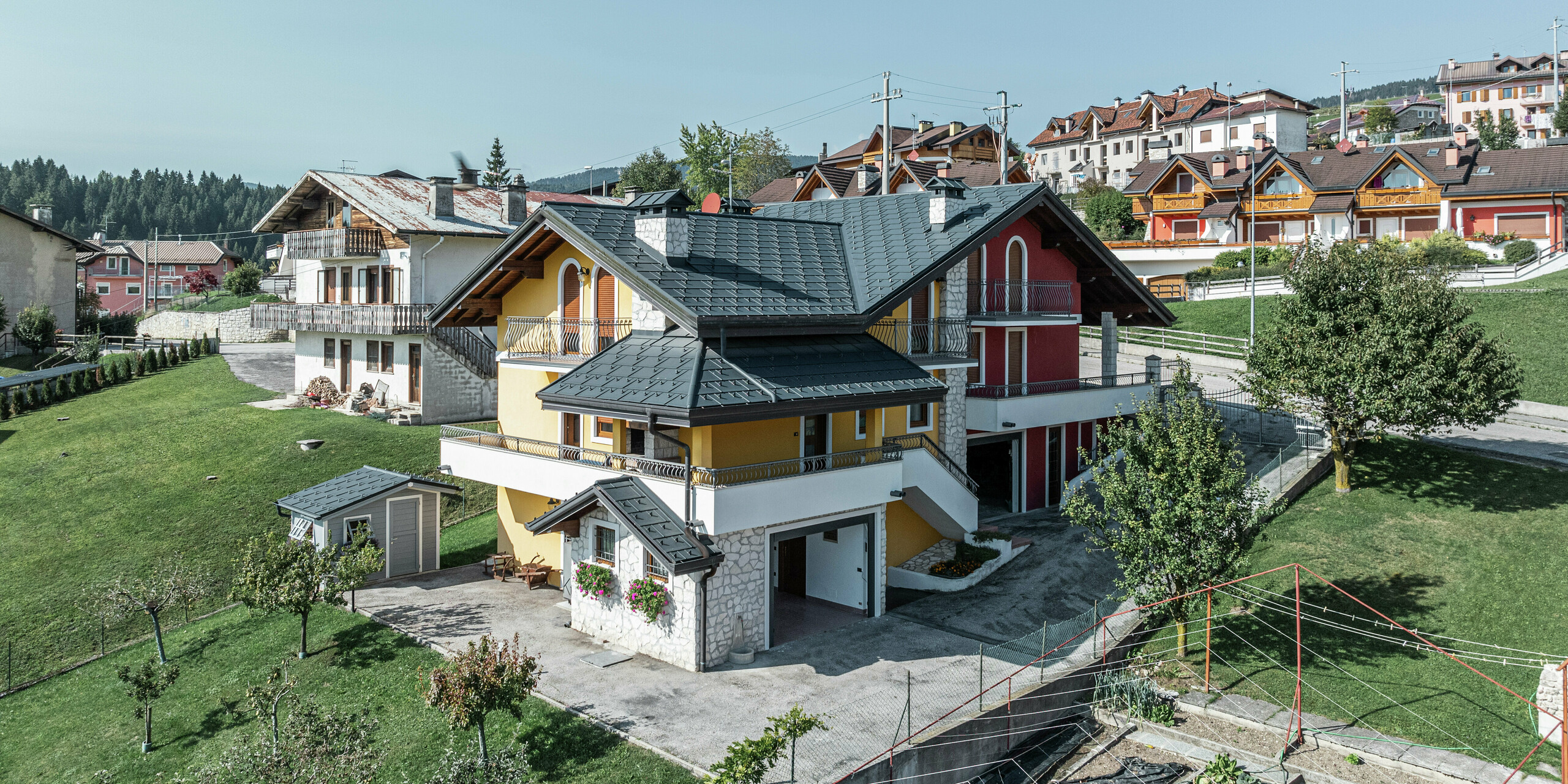 Maison individuelle à Gallio, Italie, de style alpin traditionnel avec toit PREFA moderne en P.10 gris sombre. Les panneaux de toiture PREFA R.16 offrent une protection optimale contre la neige, la pluie et le vent. La combinaison unique de façade jaune-rouge, d'éléments en pierre naturelle et d'accents en bois donne à la maison une apparence accueillante. Le système de gouttière PREFA assure un drainage fiable des précipitations. Nichée dans un paysage vallonné verdoyant, cette maison allie le charme alpin à une technologie de toiture durable et nécessitant peu d'entretien.