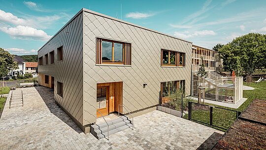 L'image présente la crèche municipale d'Alzenau, vue en plongée oblique, mettant en valeur son architecture moderne et sa remarquable façade en aluminium PREFA couleur bronze. Cette façade est composée de losanges de façade de 44 x 44 cm, créant un motif harmonieux évoquant des écailles et conférant au bâtiment un dynamisme élégant. De grandes fenêtres en bois à persiennes verticales apportent une touche chaleureuse et laissent entrer abondamment la lumière naturelle. À droite de l'image, le jardin s'ouvre sur une aire de jeux clôturée, des espaces verts et une terrasse couverte. L'alliance de l'aluminium, matériau durable et sans entretien, et de matériaux écologiques fait de ce bâtiment un exemple parfait d'architecture fonctionnelle et écologique dans le domaine de la construction éducative.