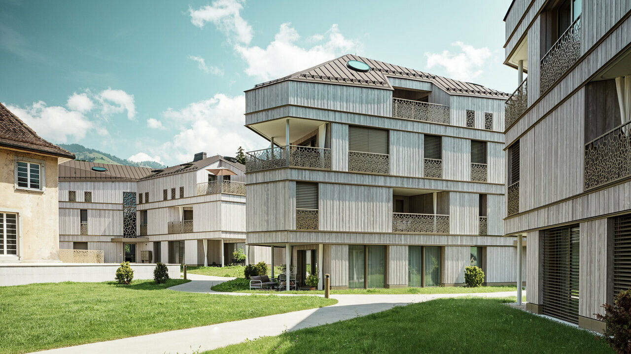  Résidence moderne à Liebwylen, Suisse, avec une architecture claire et des façades d'aspect bois. Les toits en aluminium PREFALZ de couleur brune disposent de lucarnes rondes et complètent le design contemporain. Au premier plan, un espace vert bien entretenu avec des allées sinueuses et des sièges.