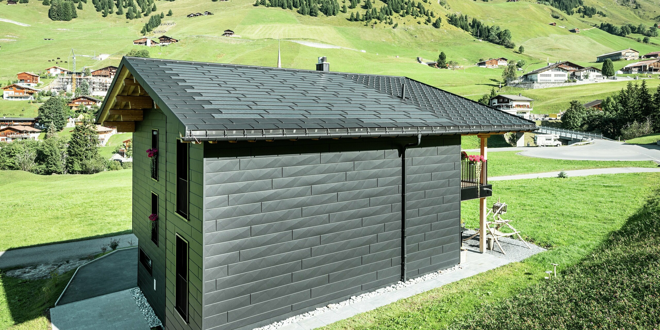 Vue latérale de la maison familiale avec une vue dégagée sur les produits de toiture et de façade PREFA en aluminium. Les tuiles solaires et les R.16 en P.10 noir offrent non seulement une protection contre les intempéries, mais également un look moderne et distinctif. La gouttière suspendue PREFA et le tuyau de descente PREFA sont parfaitement intégrés et les panneaux de façade Siding.X confèrent au bâtiment un caractère unique.