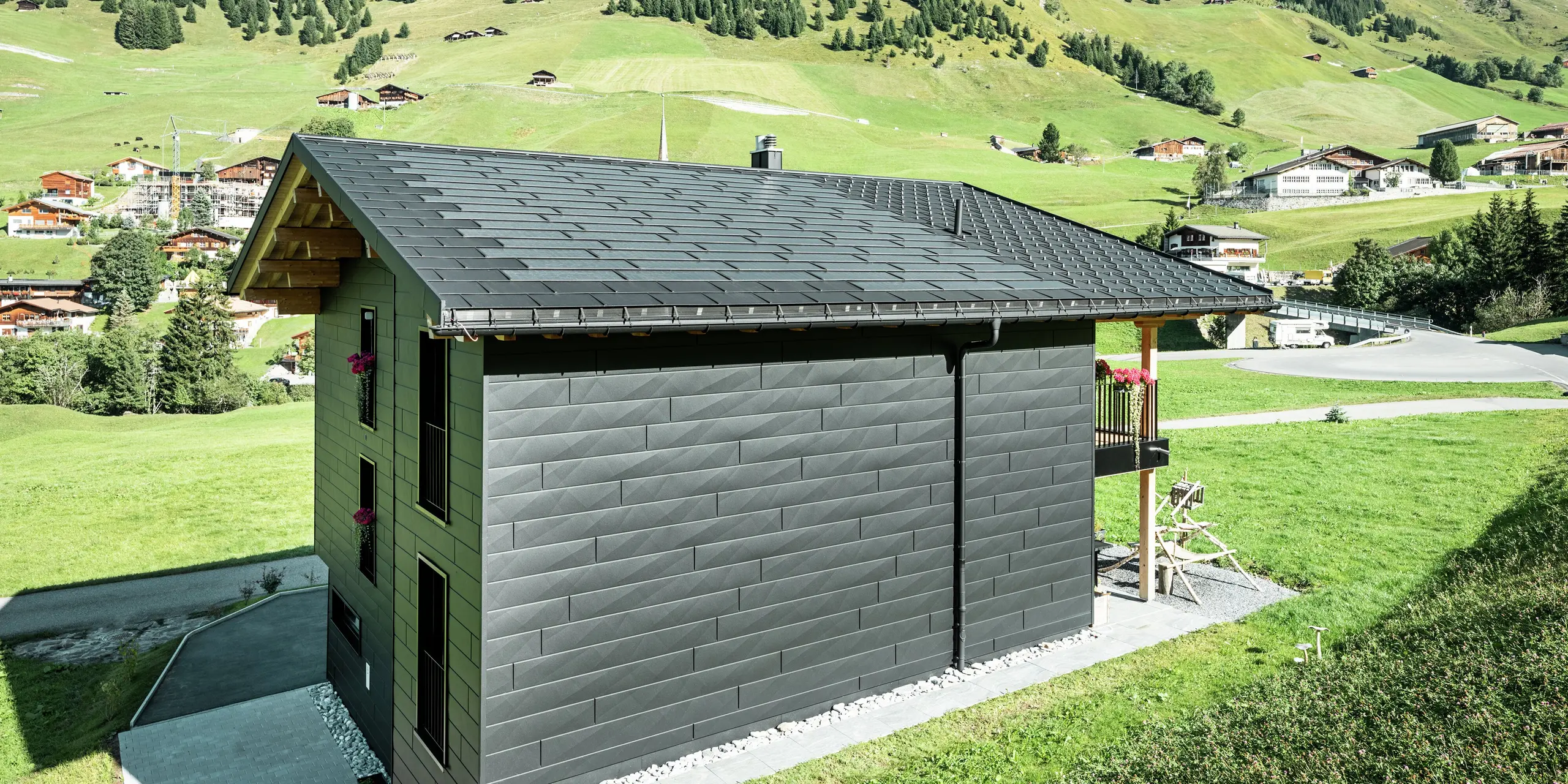 Vue latérale de la maison familiale avec une vue dégagée sur les produits de toiture et de façade PREFA en aluminium. Les tuiles solaires et les R.16 en P.10 noir offrent non seulement une protection contre les intempéries, mais également un look moderne et distinctif. La gouttière suspendue PREFA et le tuyau de descente PREFA sont parfaitement intégrés et les panneaux de façade Siding.X confèrent au bâtiment un caractère unique.