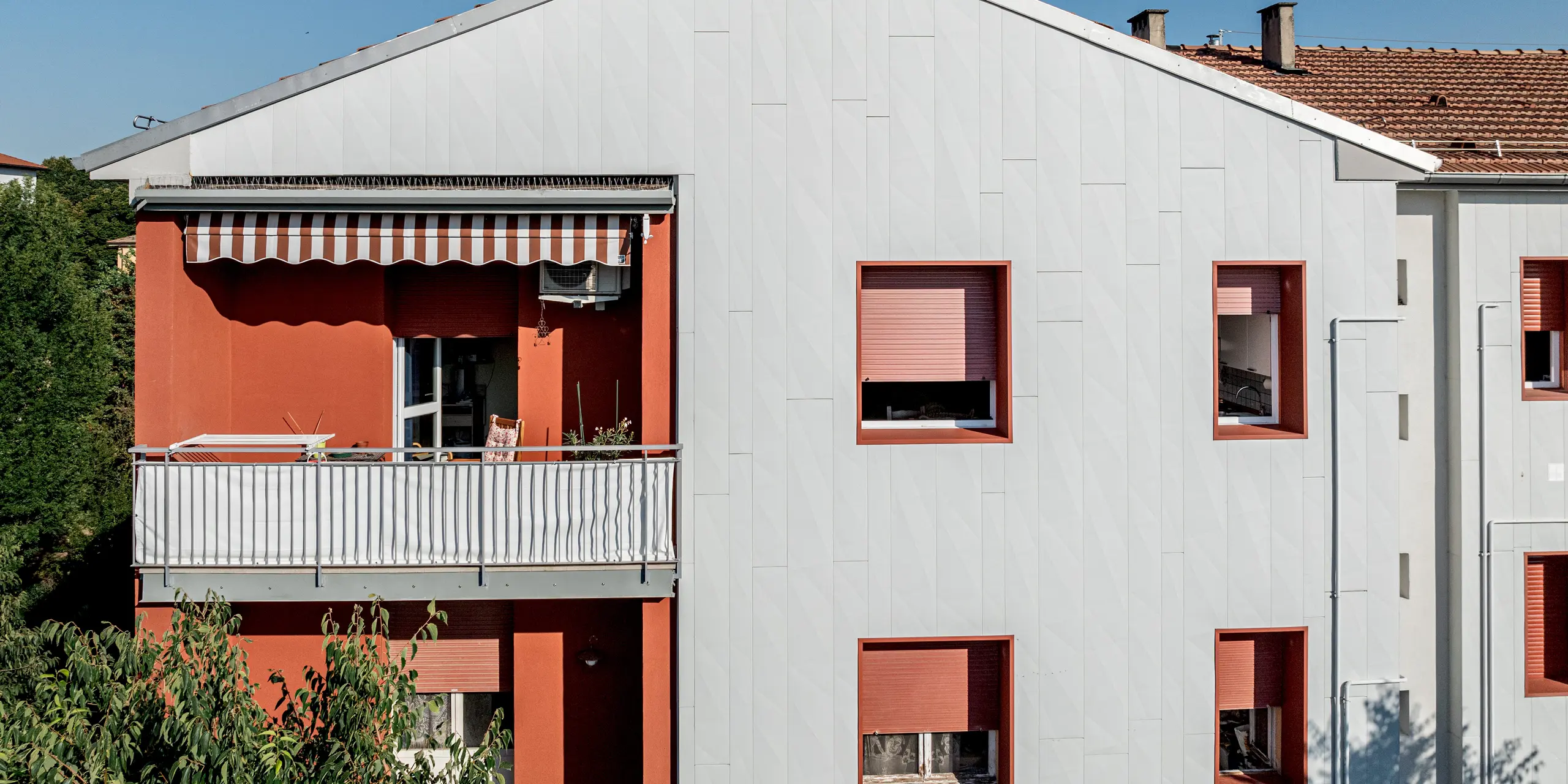 Vue détaillée du complexe résidentiel Alba B rénové, avec son pignon remarquable et sa façade à la structure affirmée, réalisée en panneaux d'aluminium PREFA Siding.X. La pose verticale des panneaux crée une texture fine et dynamique, jouant subtilement avec la lumière et l'ombre. À gauche, un balcon avec un mur enduit de rouge et un auvent apporte un contraste saisissant, tandis qu'à droite, des fenêtres profondément encastrées aux encadrements rouges soulignent le tout. La façade en aluminium PREFA impressionne par sa grande résistance aux intempéries, sa durabilité et sa facilité d'entretien, alliées à une esthétique résolument moderne.