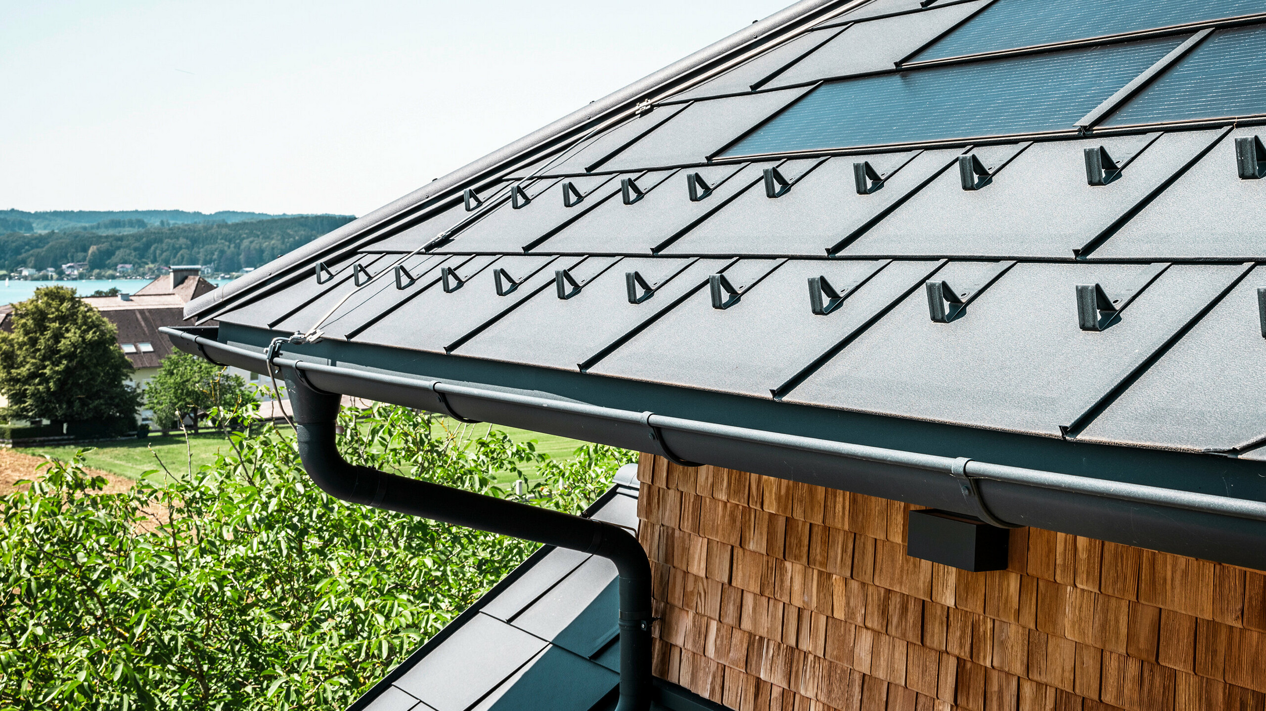 Le Zenz'n Stub'n à Schörfling am Attersee présente un toit récemment rénové avec des tuiles solaires PREFA innovantes et des R.16 PREFA durables en P.10 anthracite. Le toit élégant en aluminium allie un design moderne à une production d'énergie durable. Le système d'évacuation des eaux pluviales, composé d'un tuyau de descente et d'une gouttière suspendue en P.10 anthracite, s'intègre également harmonieusement dans l'ensemble et souligne la finition de haute qualité. Le bleu du lac Attersee scintille en arrière-plan.