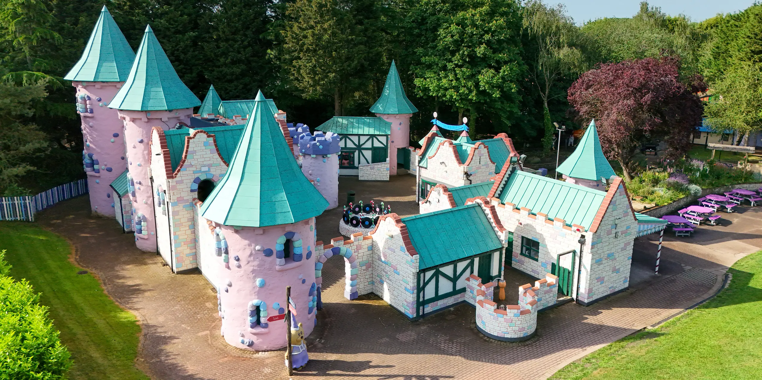 Vue de face du château féerique de Sundown Adventureland, orné de nombreuses tourelles fantaisistes, de toits coniques pointus et de façades aux teintes pastel finement détaillées. Les remarquables toitures en aluminium PREFALZ, couleur P.10 vert de gris, attirent immédiatement le regard et soulignent les formes imaginatives grâce à leurs joints debout précis. Les différentes pentes de toit et les géométries complexes sont exécutées avec élégance. Par ailleurs, les toitures PREFALZ impressionnent par leur grande durabilité, leur résistance à la corrosion et leur protection fiable – des atouts idéaux pour une architecture de loisirs d'une beauté durable.