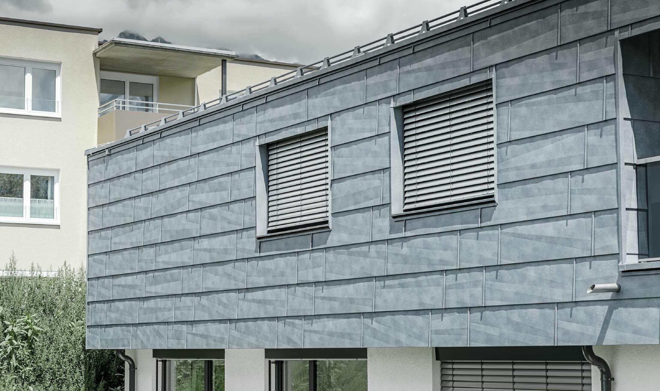Surface de façade avec deux fenêtres ; la façade est habillée avec le panneau de façade FX.12 PREFA couleur gris pierre.