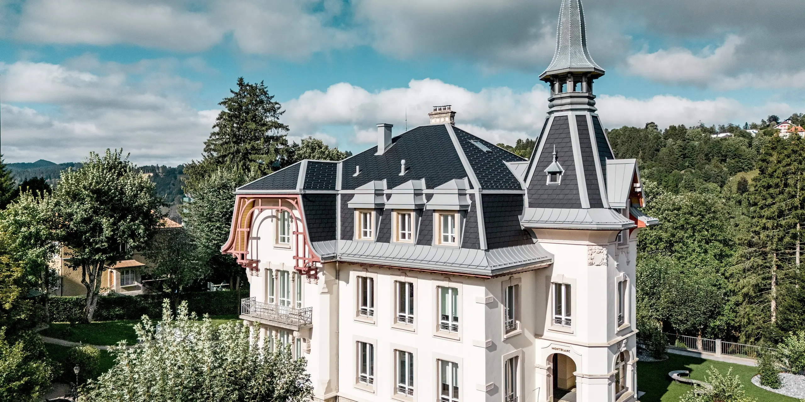 Atmosphärische Frontansicht der Manufacture Horlogère du Locle in der Schweiz mit eindrucksvollem Turm und dynamischer Dachlandschaft als Eyecatcher. Die komplexen, steil geneigten Dachflächen werden durch PREFA Dachschindeln in P.10 Schwarz präzise strukturiert, während PREFALZ in Patinagrau elegante Übergänge, Gauben und Rundungen formt. Das hochwertige Aluminiumdach überzeugt durch geringes Gewicht, hohe Widerstandsfähigkeit und extreme Langlebigkeit – eine perfekte Kombination aus architektonischer Ästhetik, technischer Präzision und nachhaltigem Schutz historischer Bausubstanz.