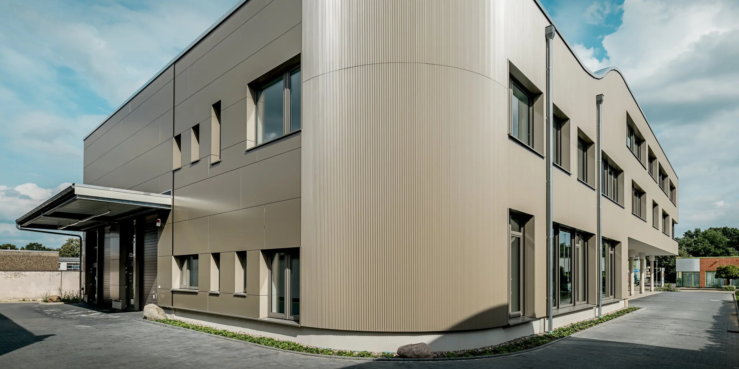 L'image présente le bâtiment Festo à Langenhagen, caractérisé par une remarquable section courbe revêtue de profils extrudés triangle PREFA. L'harmonieuse combinaison des panneaux composites en aluminium lisse et des profils triangle texturés confère à la façade une esthétique moderne et dynamique. L'angle légèrement arrondi du bâtiment est particulièrement visible, lui donnant une allure élégante, presque futuriste. Les fenêtres sont parfaitement intégrées à la façade, soulignant la pureté des lignes. L'utilisation des matériaux PREFA, durables et résistants aux intempéries, garantit une solution à la fois esthétique et adaptée à l'architecture moderne.