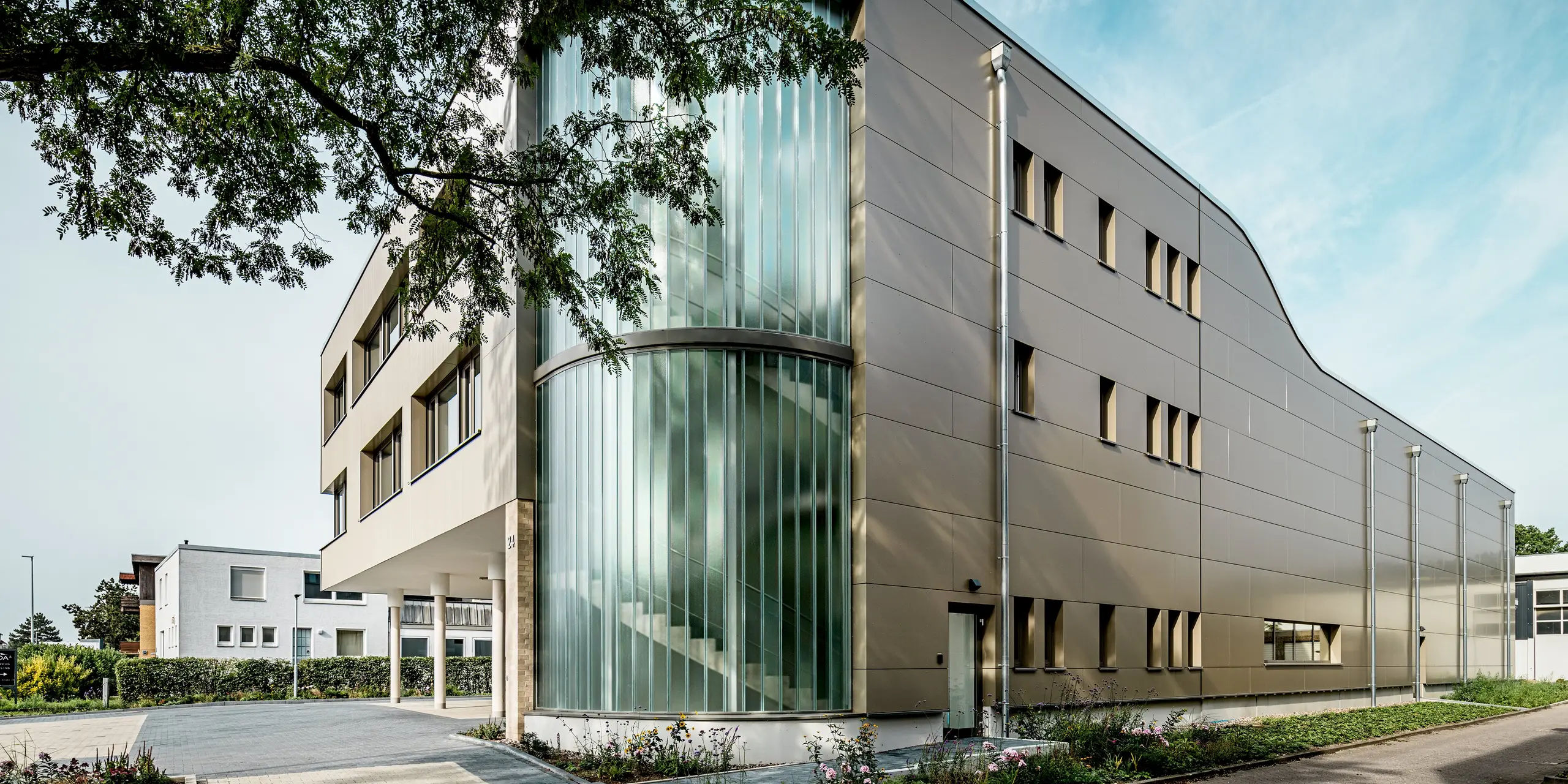 L'image présente le bâtiment Festo à Langenhagen, dont la façade remarquable est composée de panneaux composites en aluminium PREFA, dans une élégante teinte bronze. L'angle arrondi du bâtiment attire particulièrement le regard, grâce à ses surfaces vitrées verticales qui inondent l'intérieur de lumière naturelle. Cette paroi de verre crée un contraste saisissant avec la répartition uniforme des fenêtres et la pureté du design de la façade. L'utilisation des produits en aluminium PREFA garantit une solution non seulement esthétique, mais aussi durable et résistante aux intempéries. L'harmonieuse combinaison de design moderne et de fonctionnalité fait de ce bâtiment un exemple remarquable d'architecture contemporaine.
