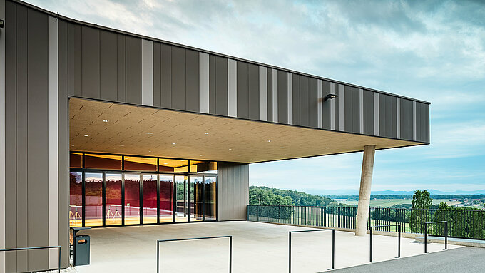 Vue extérieure du centre événementiel ZIB Nikolai à St. Veit, avec une façade moderne en aluminium dans les tons marron et beige-gris. L'entrée est couverte, soutenue par une colonne de béton inclinée, et offre une vue sur le paysage verdoyant environnant et les collines lointaines sous un ciel nuageux.