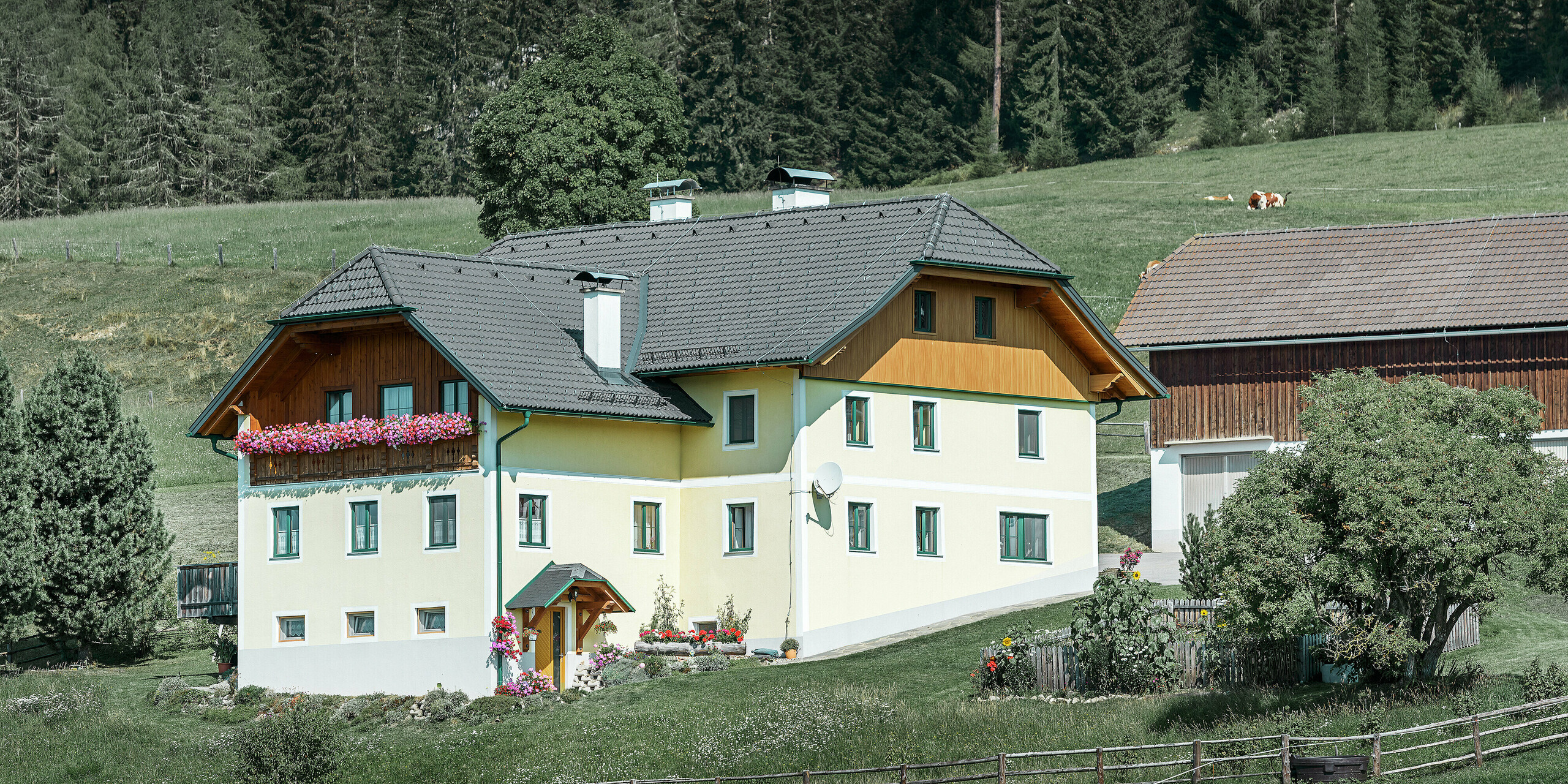 Panoramaaufnahme eines Landhauses in Ranten. Die Giebel des Hauses sind mit Holz und Aluminiumpaneelen in Holzoptik verkleidet. Der Unterschied zwischen Holz und Aluminium ist kaum zu erkennen. Die neu verkleidete Giebelseite wurde mit PREFA Sidings in Eiche natur ausgestattet. Das traditionelle Wohnhaus ist umgeben von Wiesen, Bäumen und einem angrenzenden Stadel.