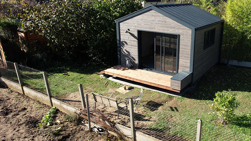 Vue de face d'un lodge situé au pied d'un jardin potager, dont la couverture a été choisie en aluminium PREFA, teinte anthracite.