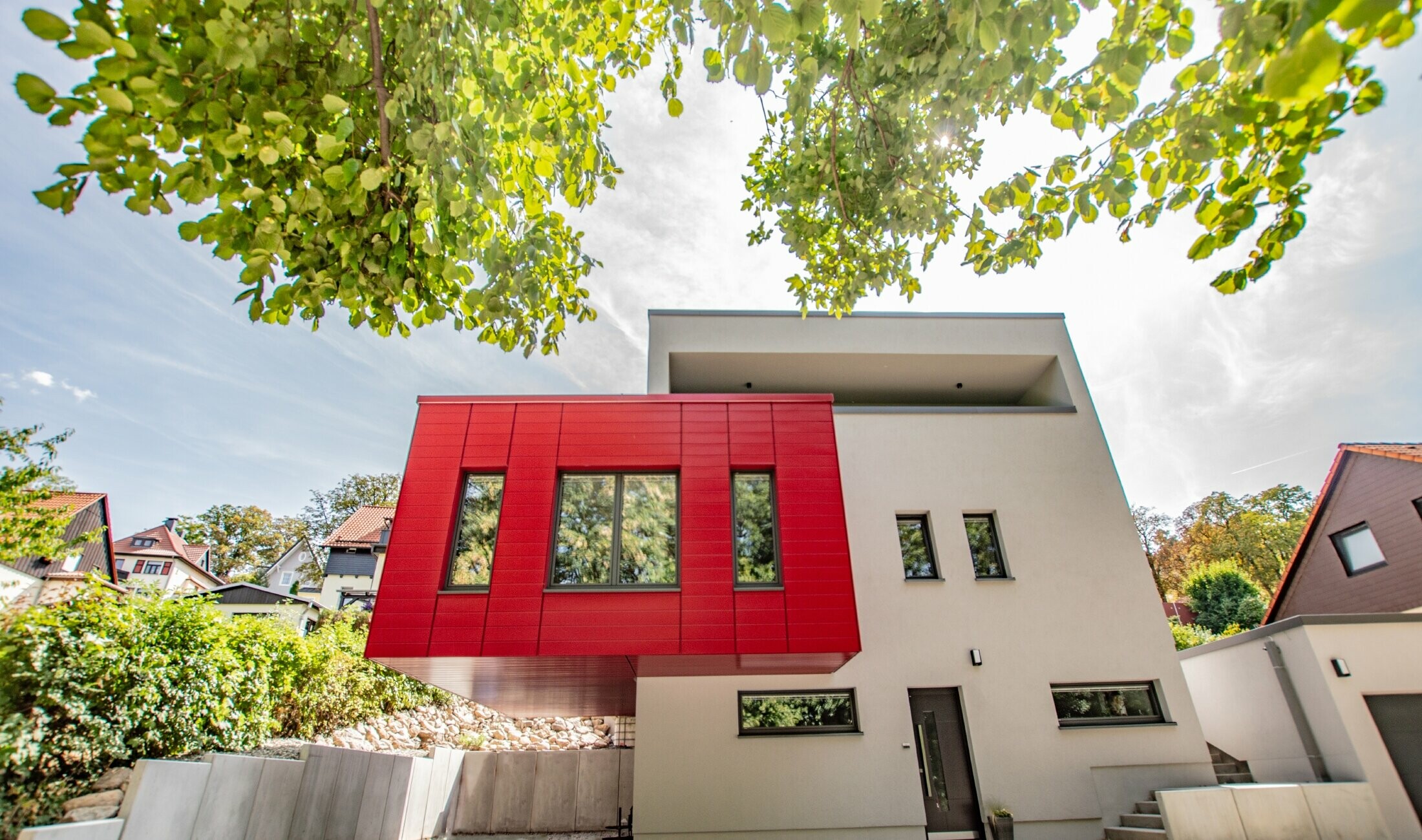 Partie de bâtiment en porte-à-faux recouverte de Sidings PREFA couleur rouge rubis. Grand oriel rouge qui détonne par rapport à la façade blanche.