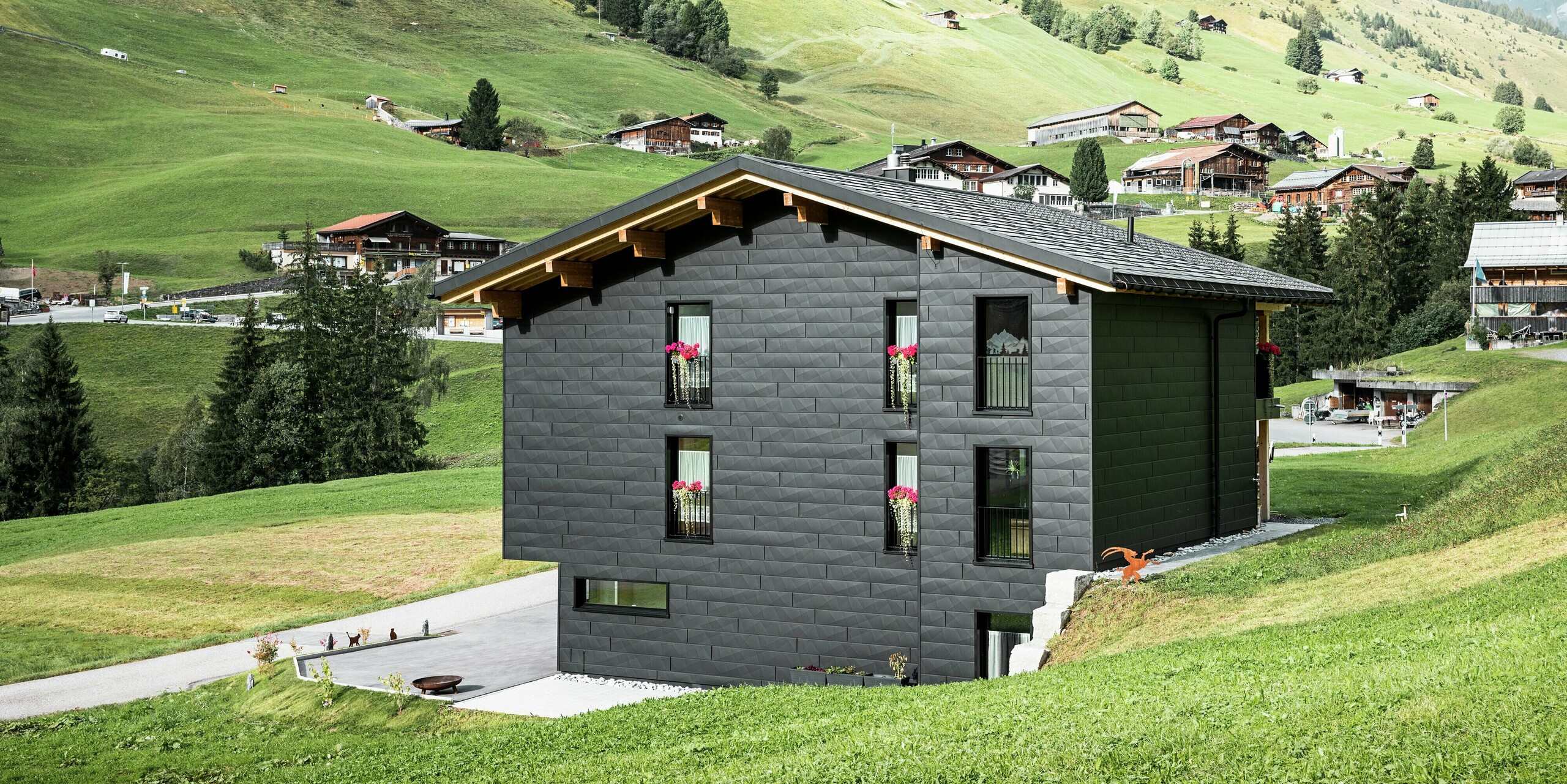 Vue arrière de la maison familiale à Ascharina, en Suisse, avec une façade en aluminium remarquable en PREFA Siding.X en P.10 noir. Les bords longitudinaux et transversaux des panneaux en aluminium confèrent au bâtiment une structure unique et moderne. Les grandes fenêtres captent beaucoup de lumière et s'harmonisent avec les éléments en bois naturel qui accentuent la façade.