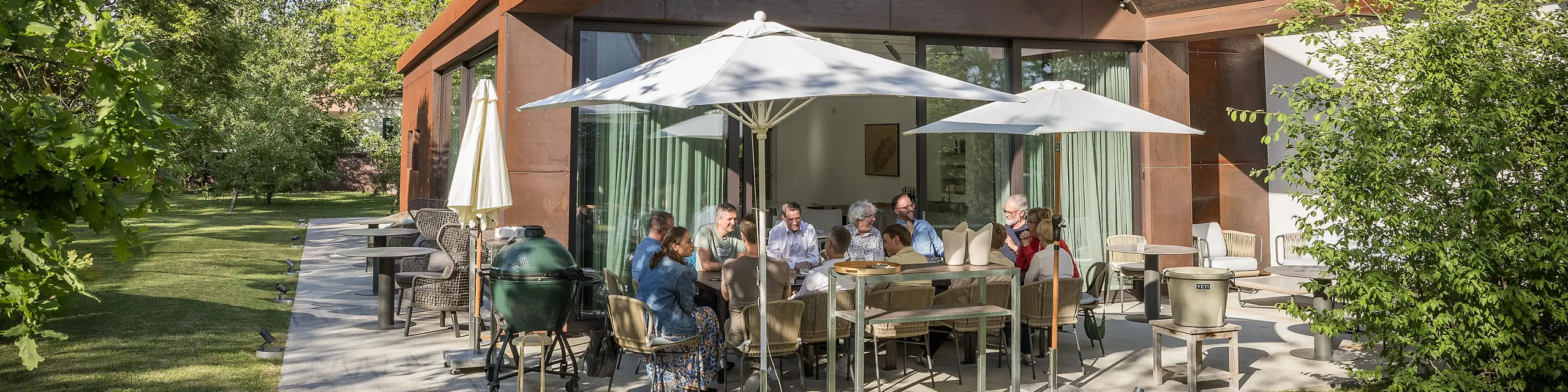 PREFARENZEN 2027 : Dialogue d’experts sur la terrasse d’une résidence de vigneron moderne : les membres du jury, réunis autour d’une grande table, discutent longuement de projets architecturaux et feuillettent des ouvrages d’architecture. La scène, baignée de lumière naturelle et agrémentée d’un jardin verdoyant, laisse entrevoir les produits en aluminium PREFA sur le toit et la façade du bâtiment. L’image traduit un engagement créatif, une analyse approfondie et des échanges stimulants. Les toitures et façades PREFA incarnent des solutions innovantes, durables et d’une grande élégance, alliant design, fonctionnalité et inspiration dans les projets architecturaux.