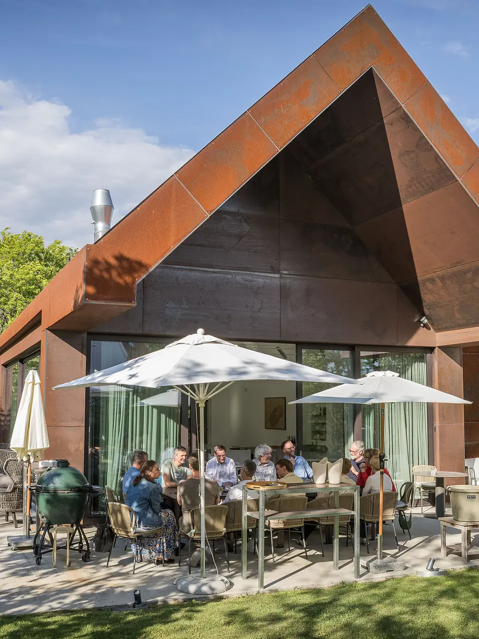PREFARENZEN 2027 : Dialogue d’experts sur la terrasse d’une résidence de vigneron moderne : les membres du jury, réunis autour d’une grande table, discutent longuement de projets architecturaux et feuillettent des ouvrages d’architecture. La scène, baignée de lumière naturelle et agrémentée d’un jardin verdoyant, laisse entrevoir les produits en aluminium PREFA sur le toit et la façade du bâtiment. L’image traduit un engagement créatif, une analyse approfondie et des échanges stimulants. Les toitures et façades PREFA incarnent des solutions innovantes, durables et d’une grande élégance, alliant design, fonctionnalité et inspiration dans les projets architecturaux.