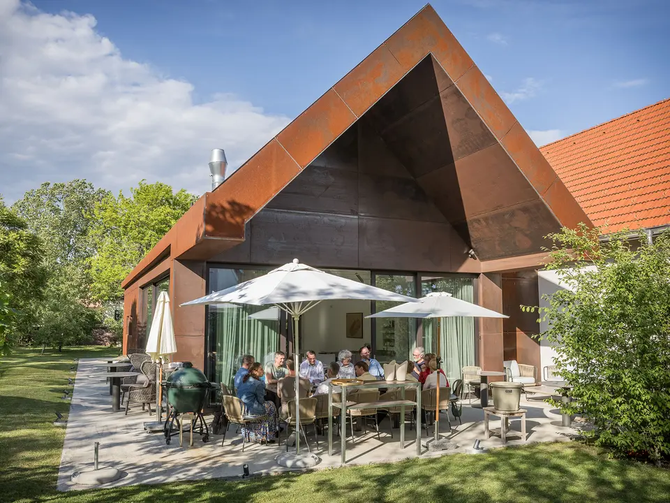 PREFARENZEN 2027 : Dialogue d’experts sur la terrasse d’une résidence de vigneron moderne : les membres du jury, réunis autour d’une grande table, discutent longuement de projets architecturaux et feuillettent des ouvrages d’architecture. La scène, baignée de lumière naturelle et agrémentée d’un jardin verdoyant, laisse entrevoir les produits en aluminium PREFA sur le toit et la façade du bâtiment. L’image traduit un engagement créatif, une analyse approfondie et des échanges stimulants. Les toitures et façades PREFA incarnent des solutions innovantes, durables et d’une grande élégance, alliant design, fonctionnalité et inspiration dans les projets architecturaux.